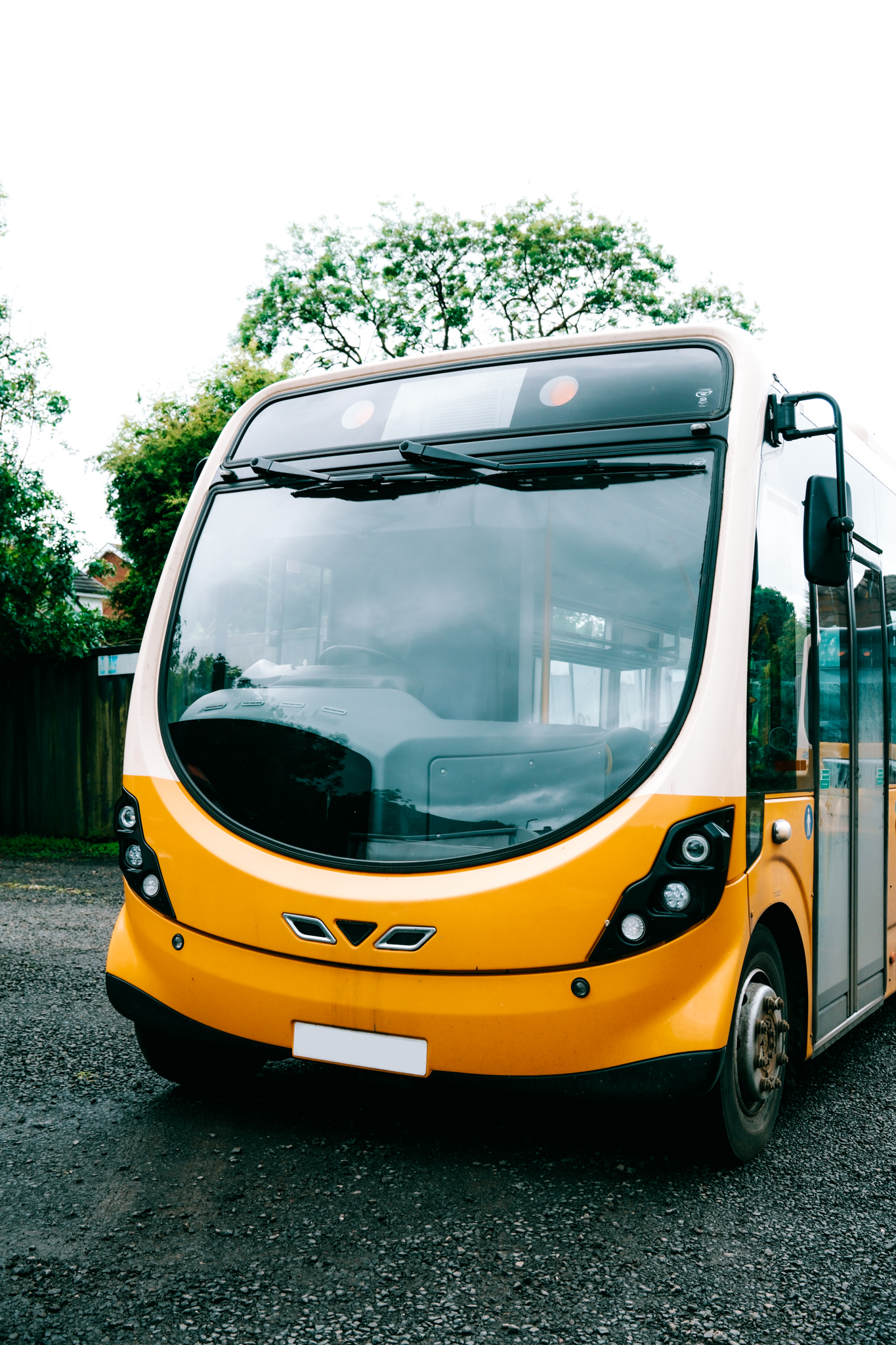 Electric yellow and white shuttle bus on a gravel lot with green trees in the background.