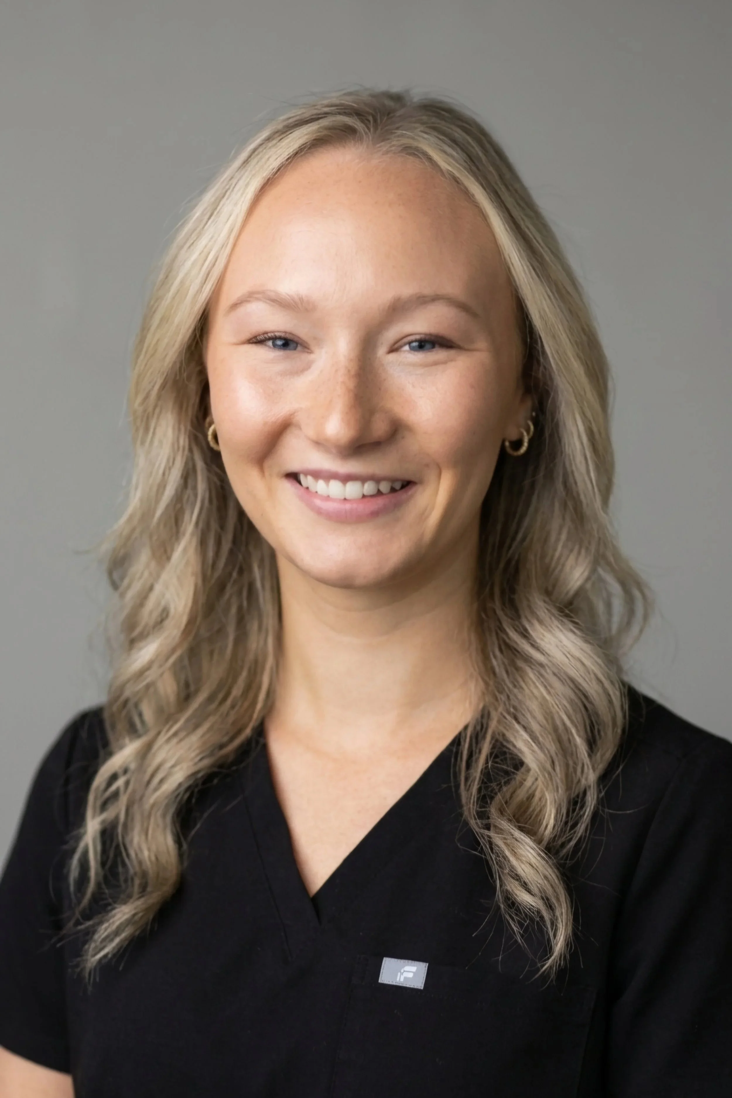 A woman with long, wavy blonde hair, blue eyes, and a light complexion, smiling, wearing a black top with a small logo, and gold hoop earrings, against a plain gray background.