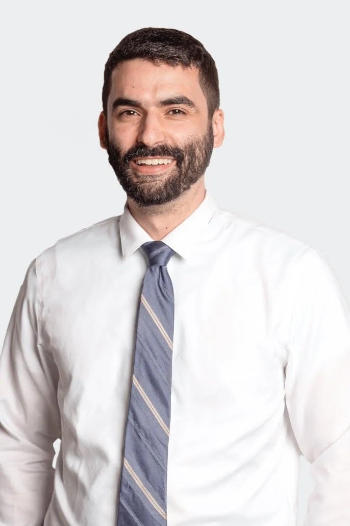 A man with dark hair and a beard wearing a white dress shirt and a blue striped tie, smiling against a plain light background.