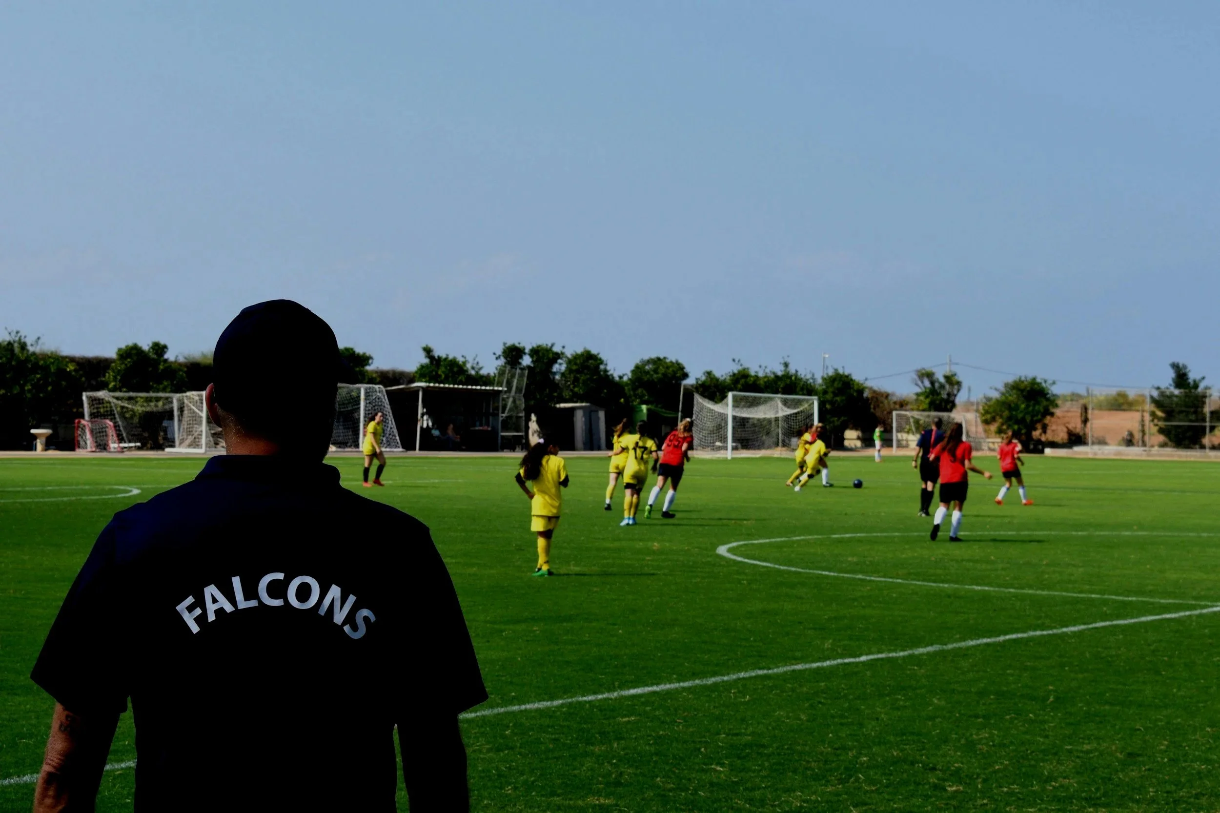 A soccer game between girls on a green field, with players in yellow and red uniforms, and a coach or spectator in the foreground wearing a black shirt with 'FALCONS' written on the back.