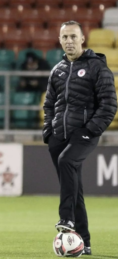 A man standing on a soccer field with his foot on a soccer ball. He is wearing a black jacket and black pants, with a serious expression. The background shows stadium seating with yellow, green, and red seats.