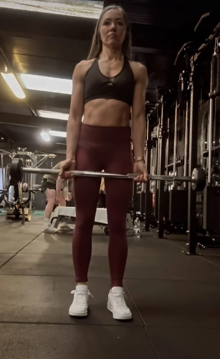 A woman in workout attire holding a barbell in a gym, with gym equipment and other people in the background.