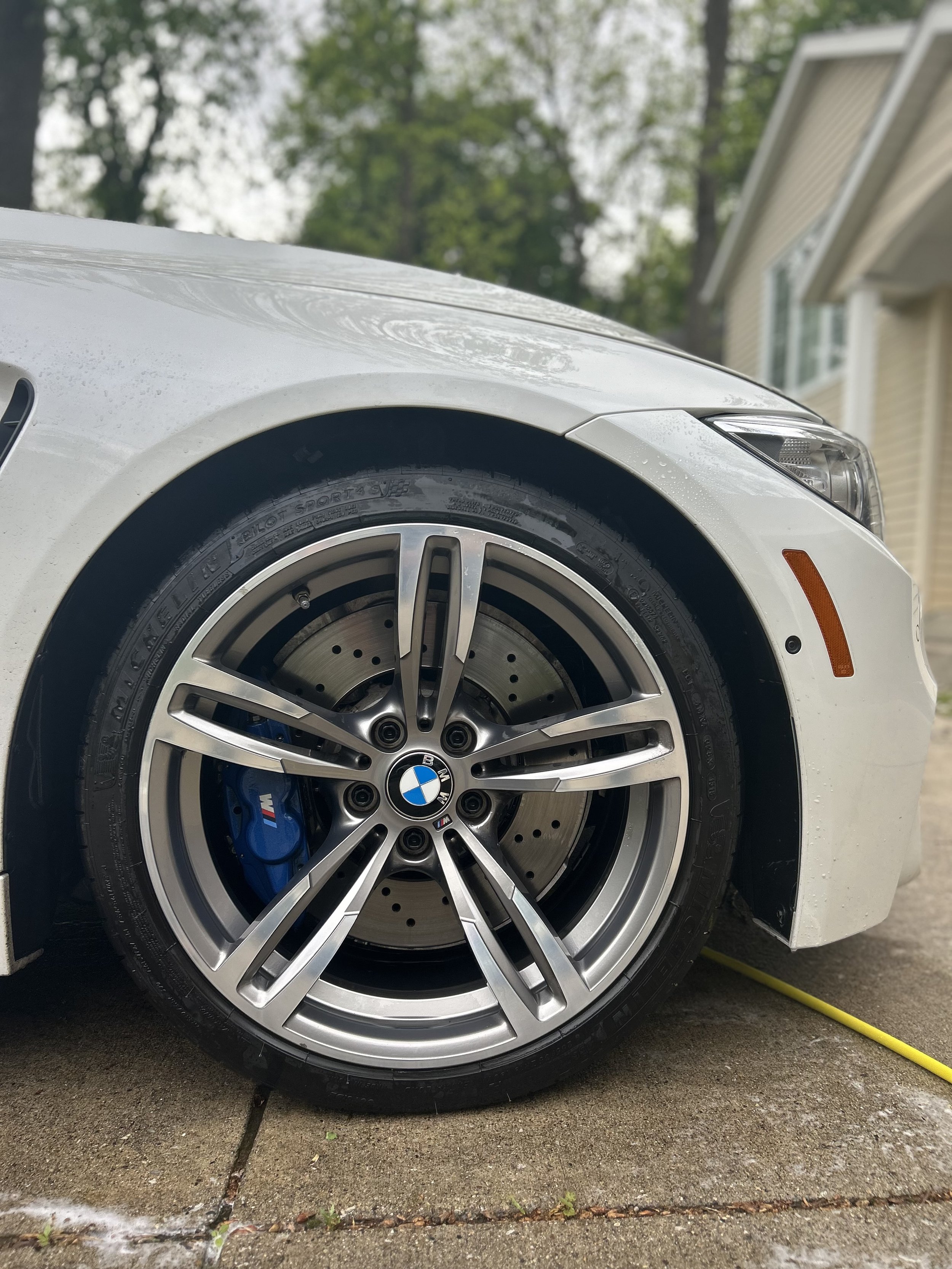 Close-up of the front wheel of a white BMW car, showing a silver rim, blue brake caliper, and perforated brake disc, parked on a wet concrete driveway.