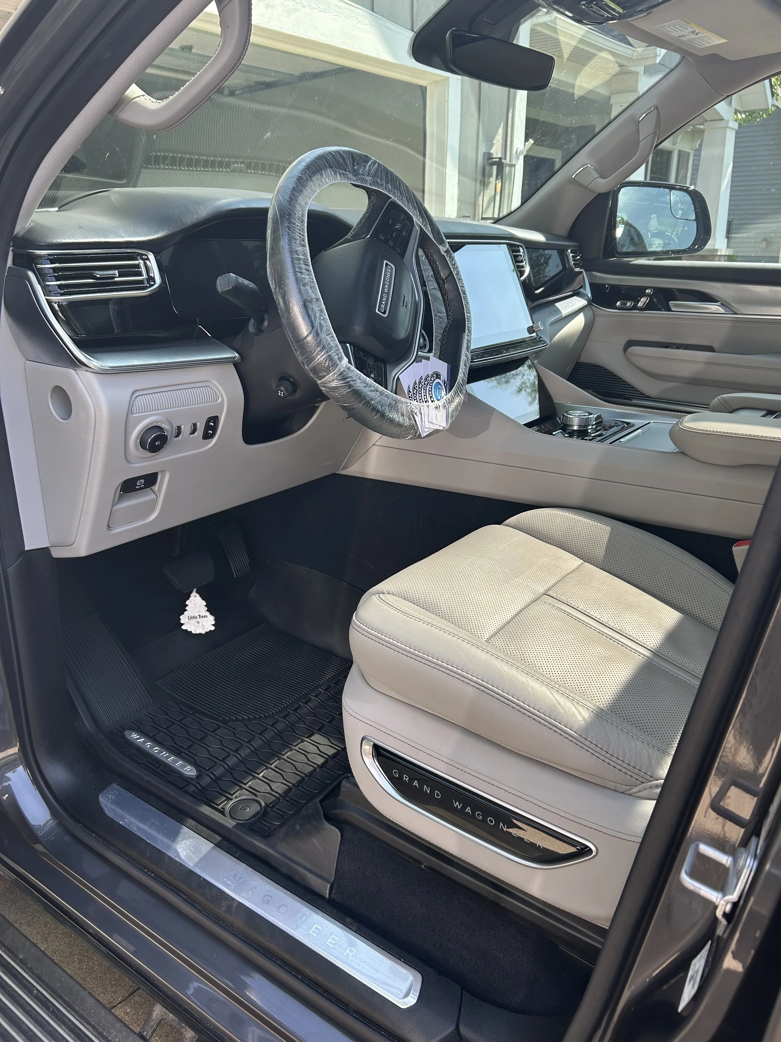 Interior of a Land Rover Discovery Grand Wagoneer SUV, showing the steering wheel with a plastic cover, a large touchscreen dashboard, and a beige leather seat with a 'Grand Wagoneer' badge.