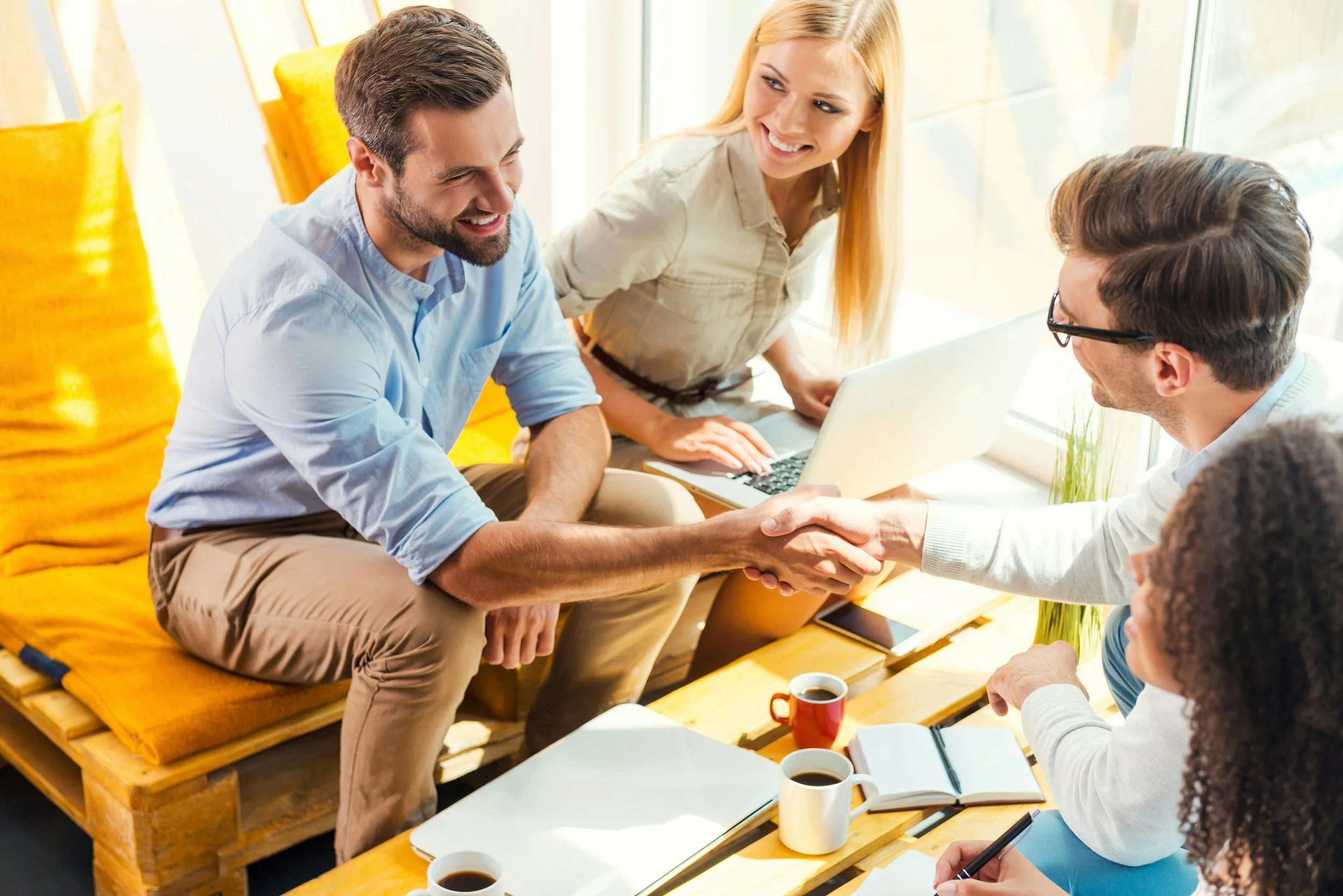 Two men shaking hands in a meeting room with a woman standing behind them, a second man and woman seated at the table, coffee cups, notebooks, and a laptop on the table.