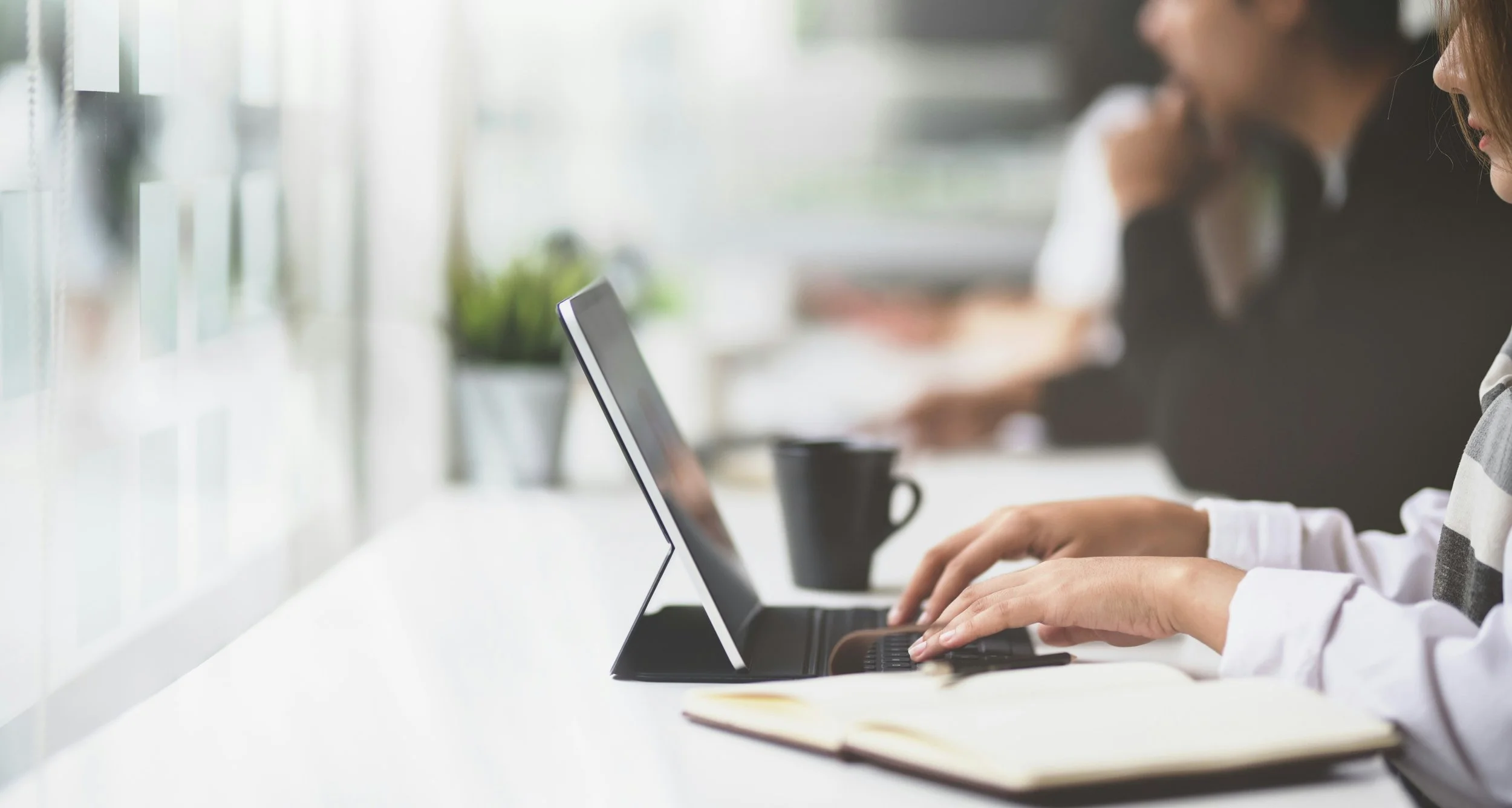 People working on laptops at a bright, modern workspace with notebooks, coffee cups, and plants.