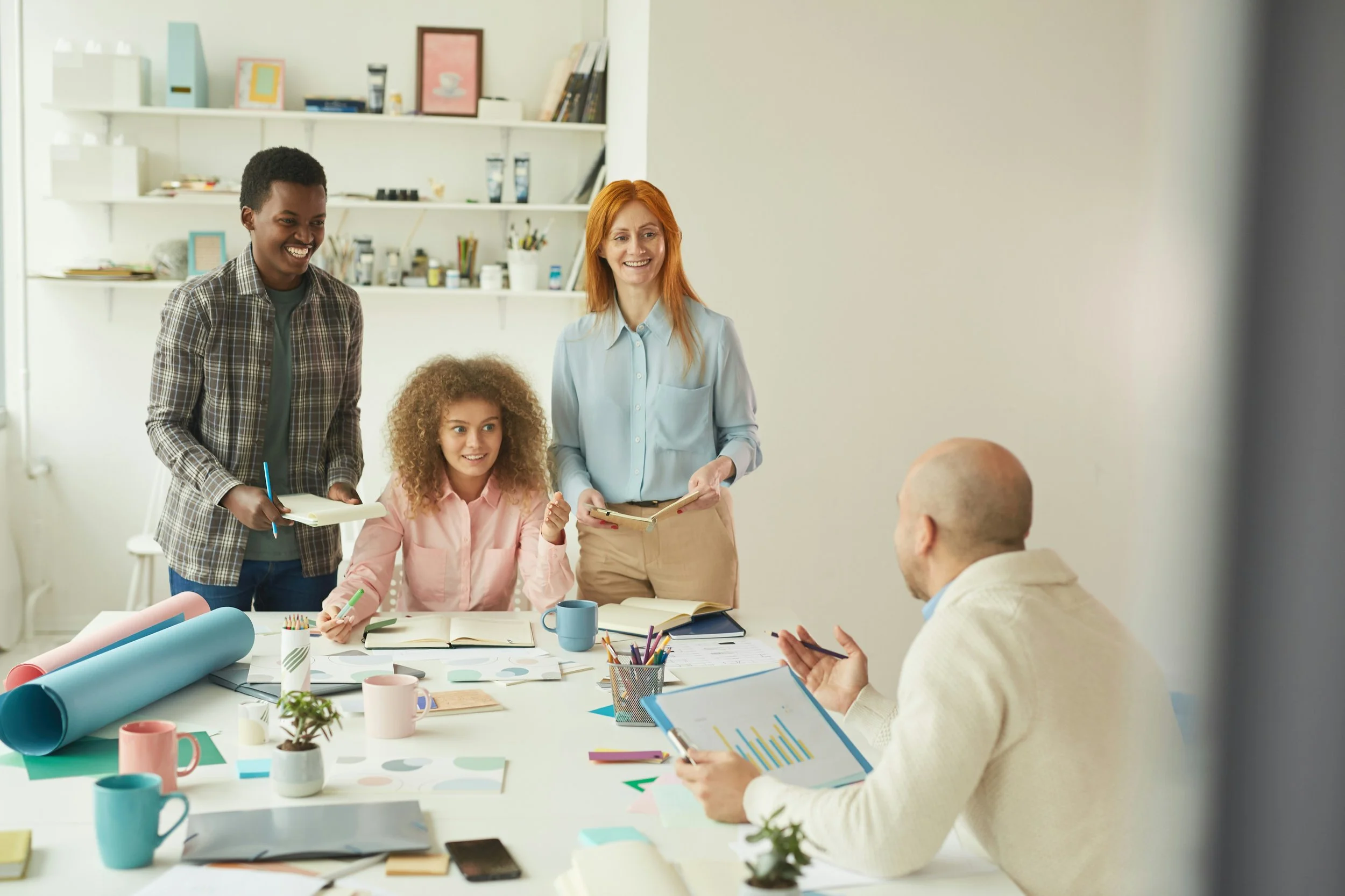 A group of four people having a meeting in a bright office. Two women and one man are standing, and one woman is seated at a table with various office supplies, papers, and coffee mugs. They are smiling and engaged in conversation.