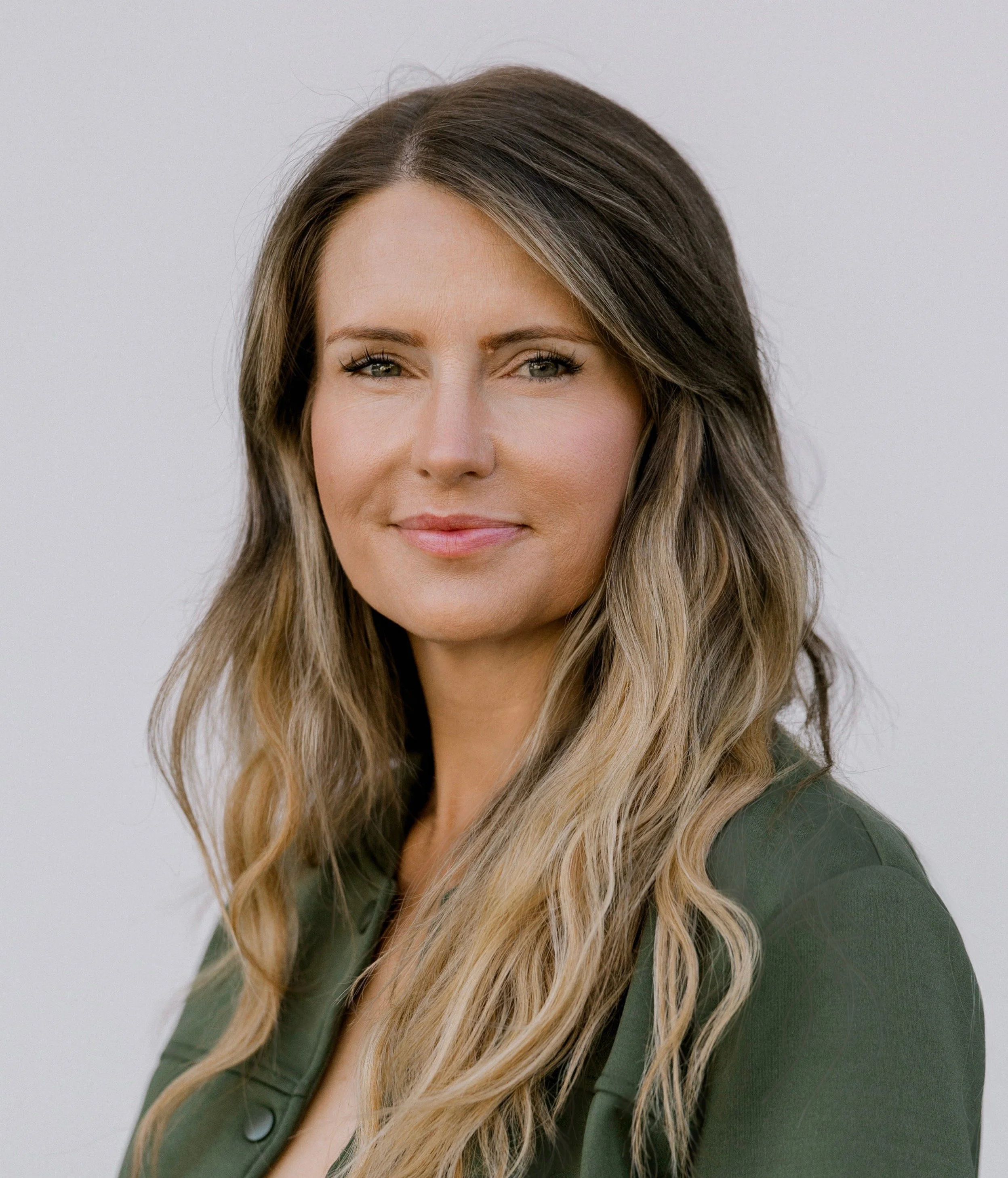 Portrait of a woman with long wavy blond hair, wearing a dark green jacket, smiling softly, against a plain light gray background.