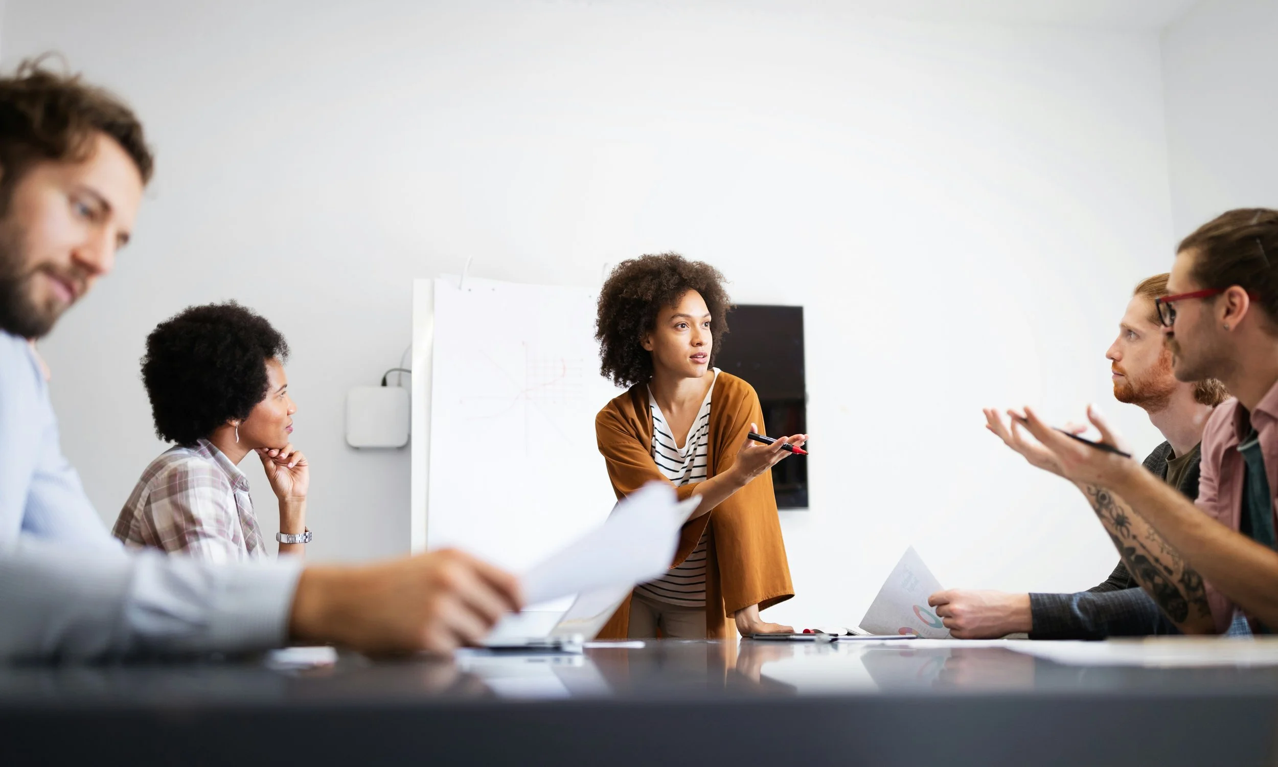 A woman leader with curly hair presenting to a diverse group of colleagues in a meeting room.
