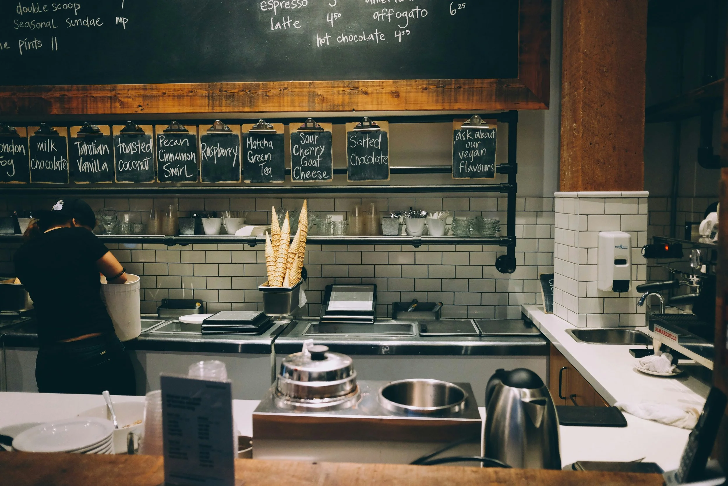 Interior of a cafe kitchen with a chalkboard menu and handwritten chalkboard signs listing flavors like matcha green tea and salted chocolate. A barista is preparing a beverage, with ice cream cones in a container on the counter. Various utensils, equipment, and a sink are visible.