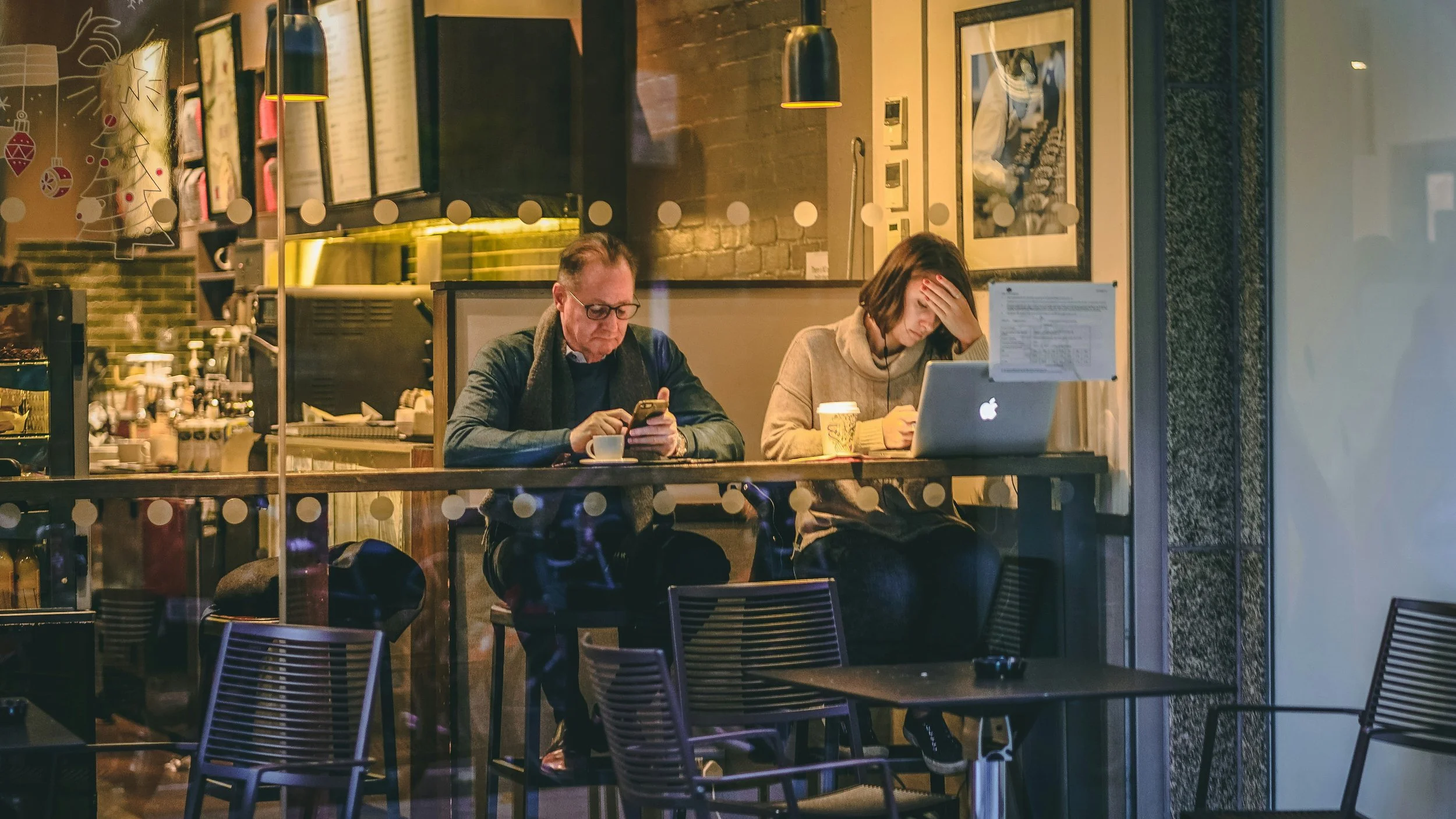 Two people sitting at a coffee shop window inside, looking at their devices, with coffee cups on the table, during the evening.
