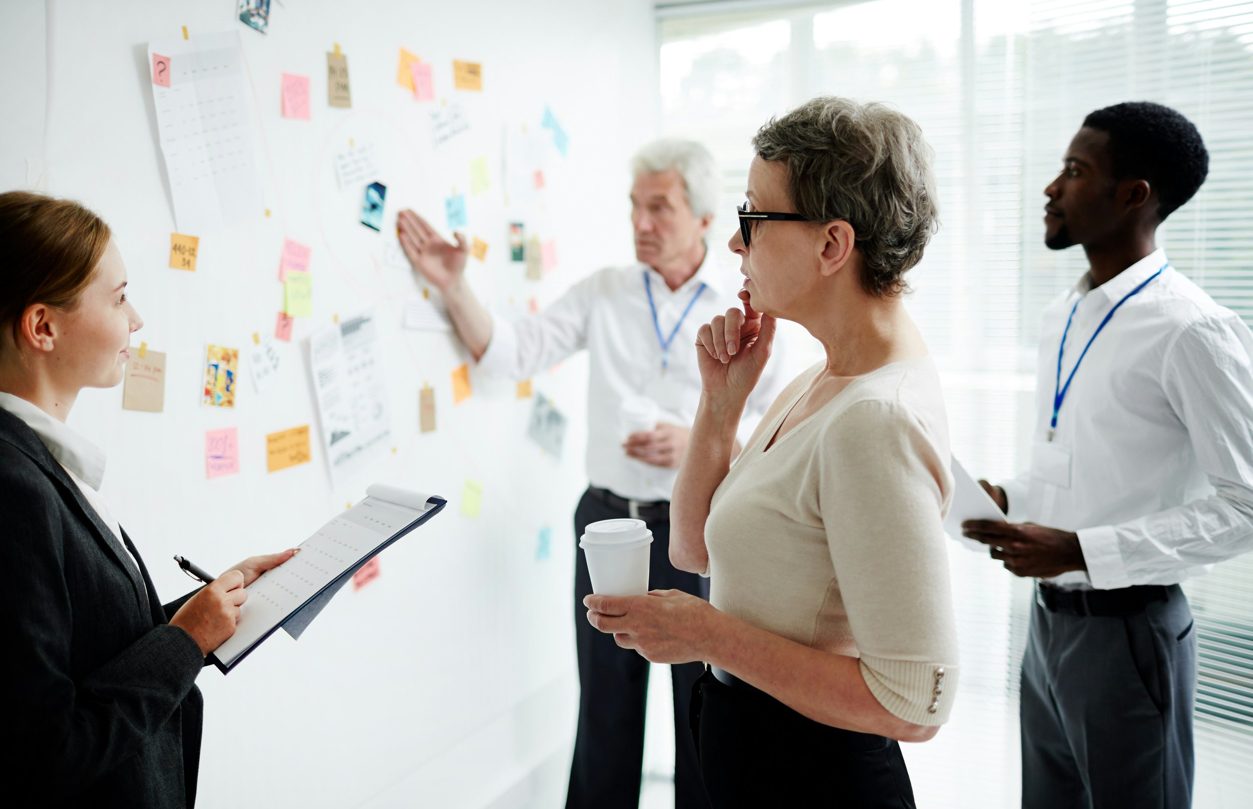 Four professionals in a meeting room engaging with a wall covered in colorful sticky notes, papers, and sketches. One woman with glasses and a coffee cup appears to be pondering, while another woman with a notepad listens. Two men, one pointing at the wall and the other holding a notebook, are explaining and listening.