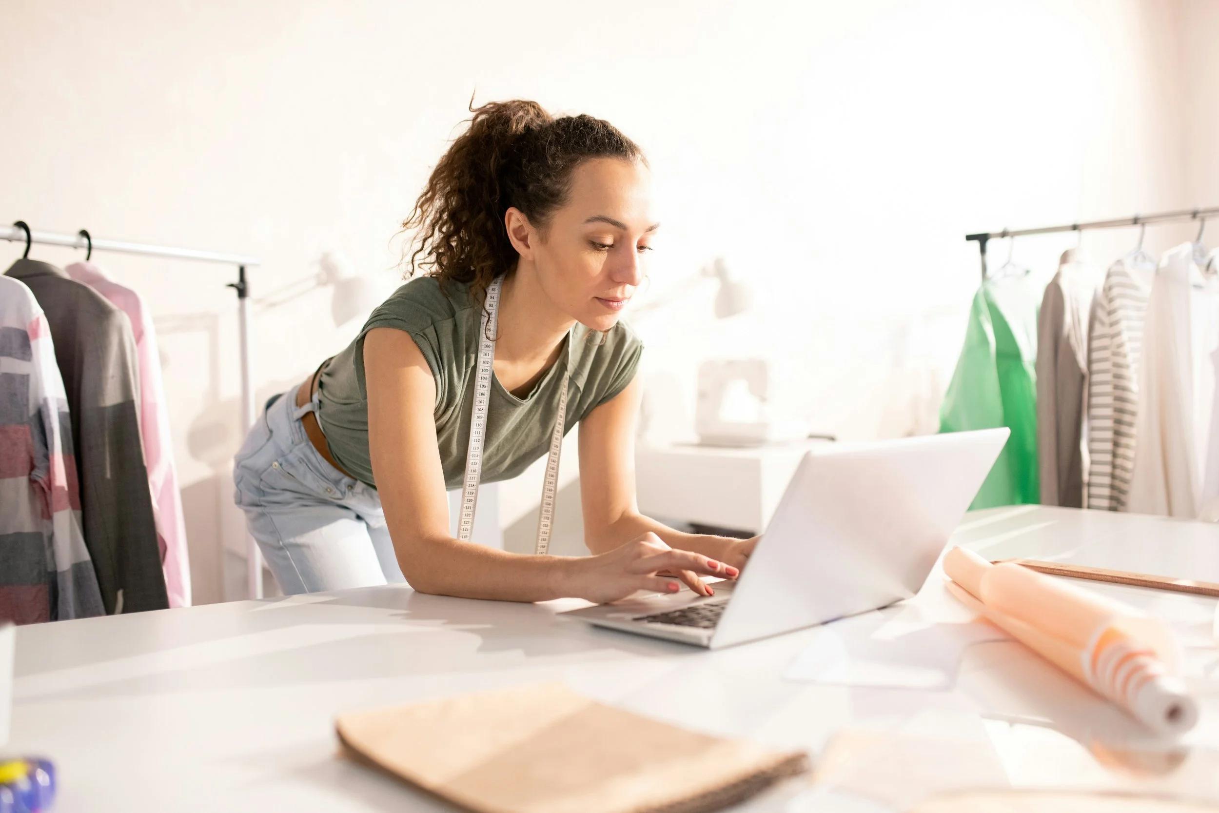Young woman in a green t-shirt leaning over a white table, working on a laptop in a fashion design studio, with clothing racks and fabric rolls visible in the background.