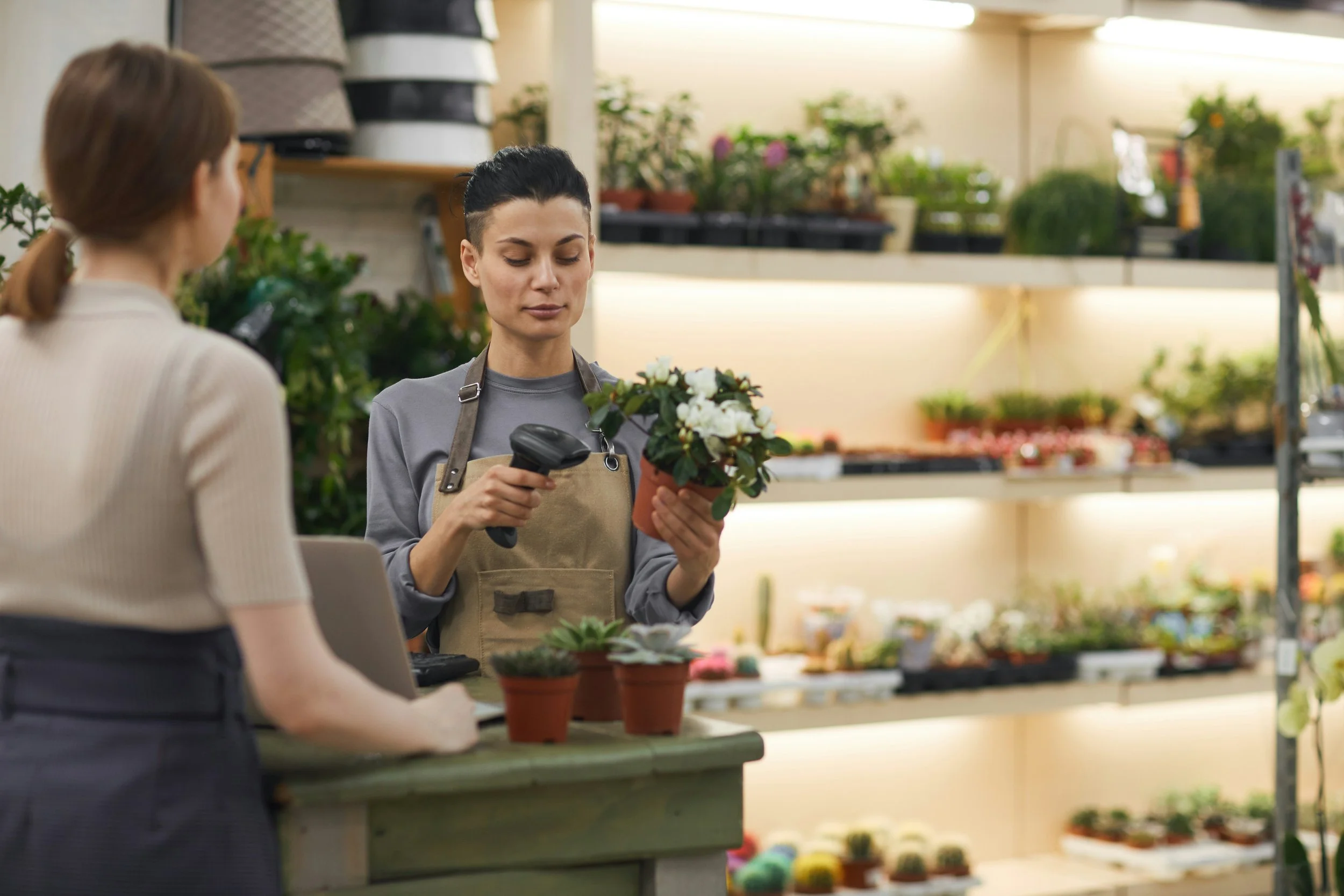 A woman with slicked-back dark hair wearing an apron checks a potted plant with a barcode scanner in a plant shop, while another woman with light brown hair in a ponytail looks on. The background is filled with shelves of plants and gardening supplies.