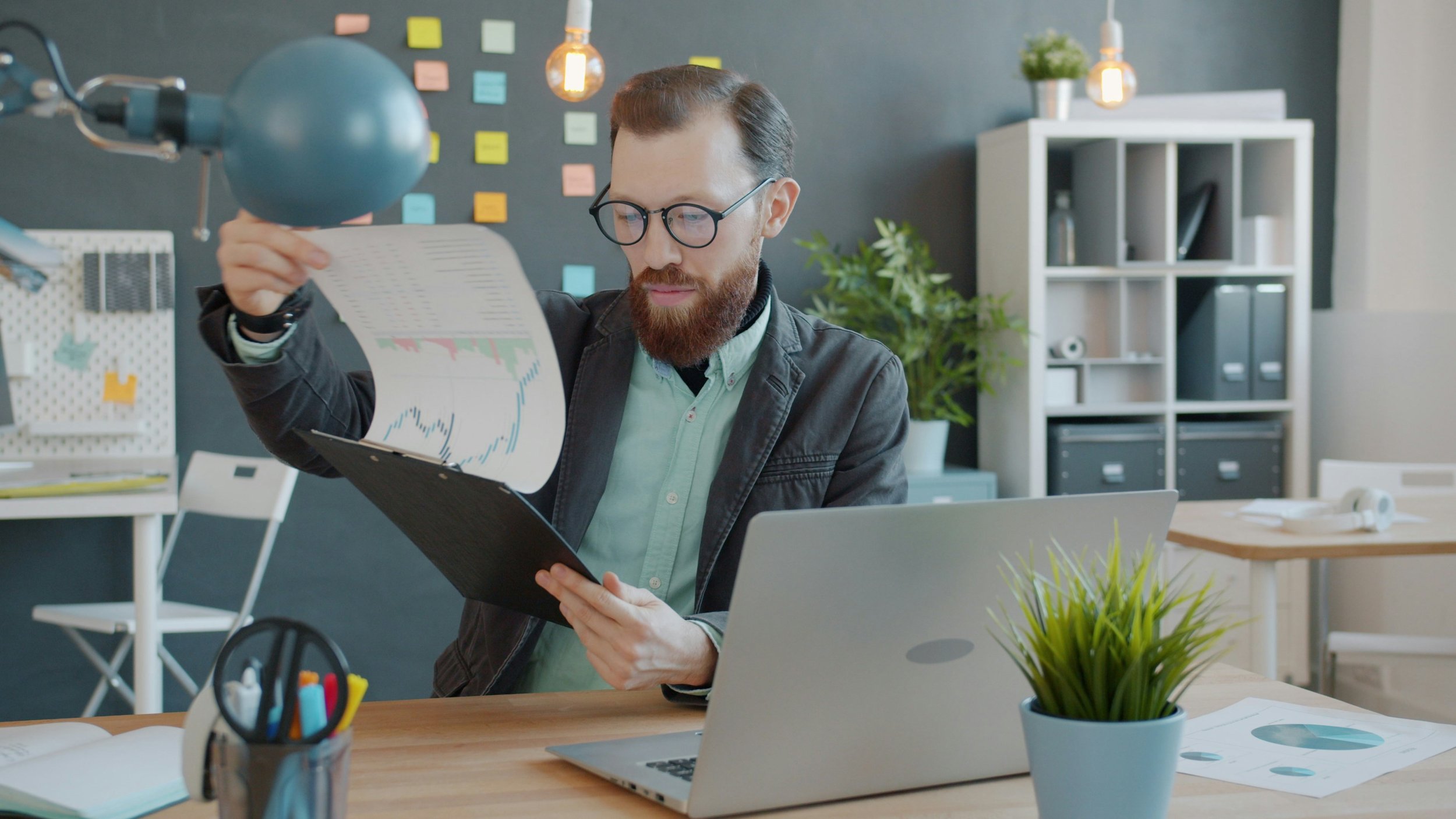 Man with glasses and beard reviewing financial charts at his desk in an office.