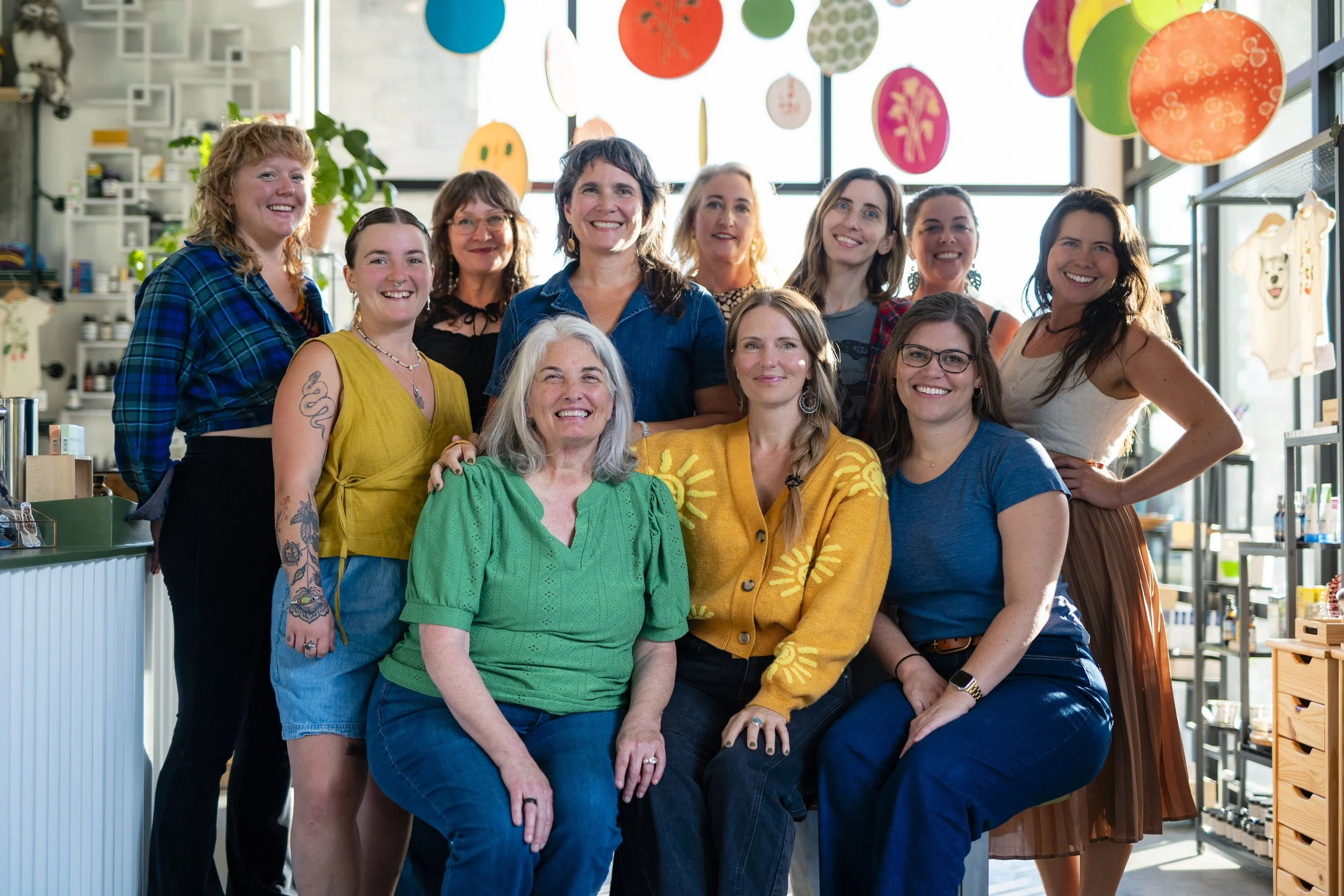 A group of 12 women smiling for a photo in a brightly decorated room with colorful hanging ornaments and large windows.