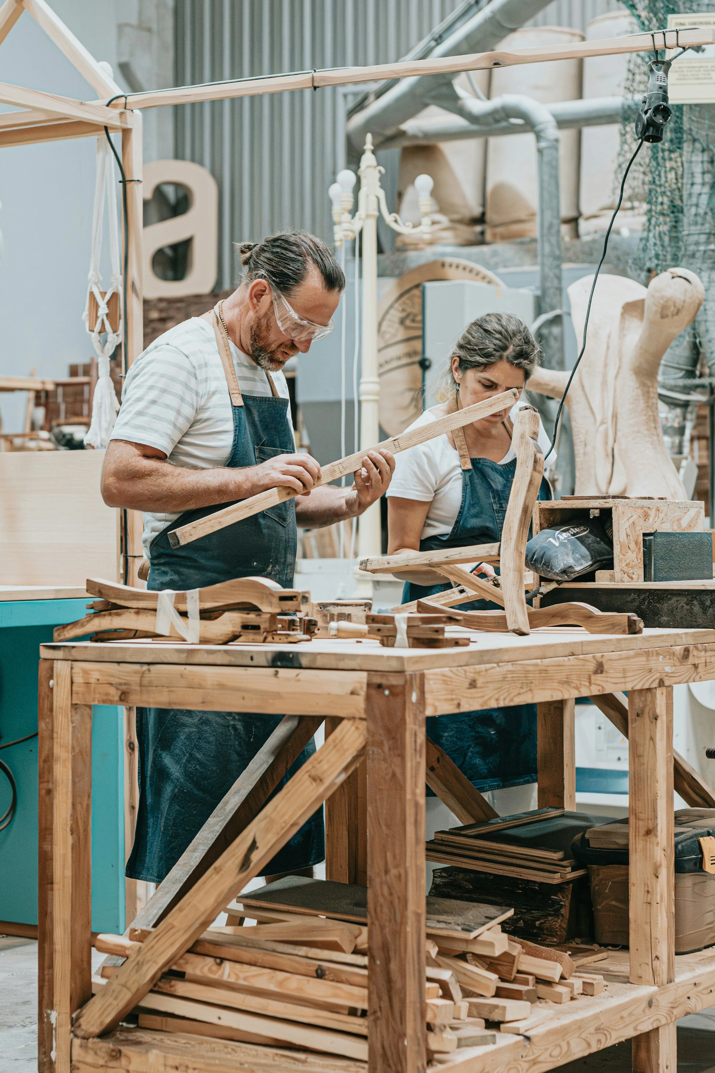 Two people working on woodworking projects at a workshop, surrounded by wooden pieces, tools, and various wood sculptures in the background.