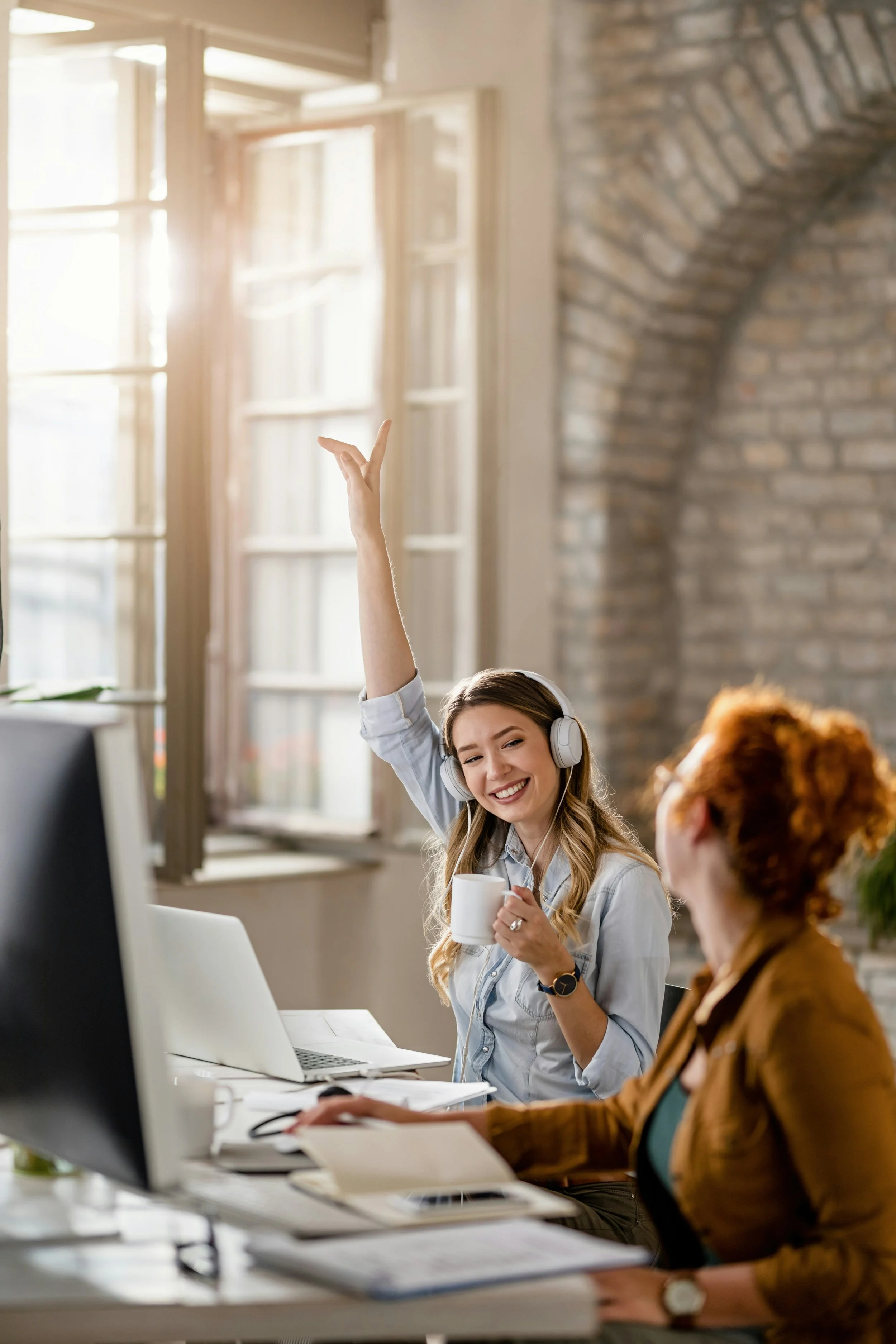 A woman wearing headphones, smiling, holding a mug, and raising her hand in an office with sunlight coming through open windows, while another woman is seated at the desk.