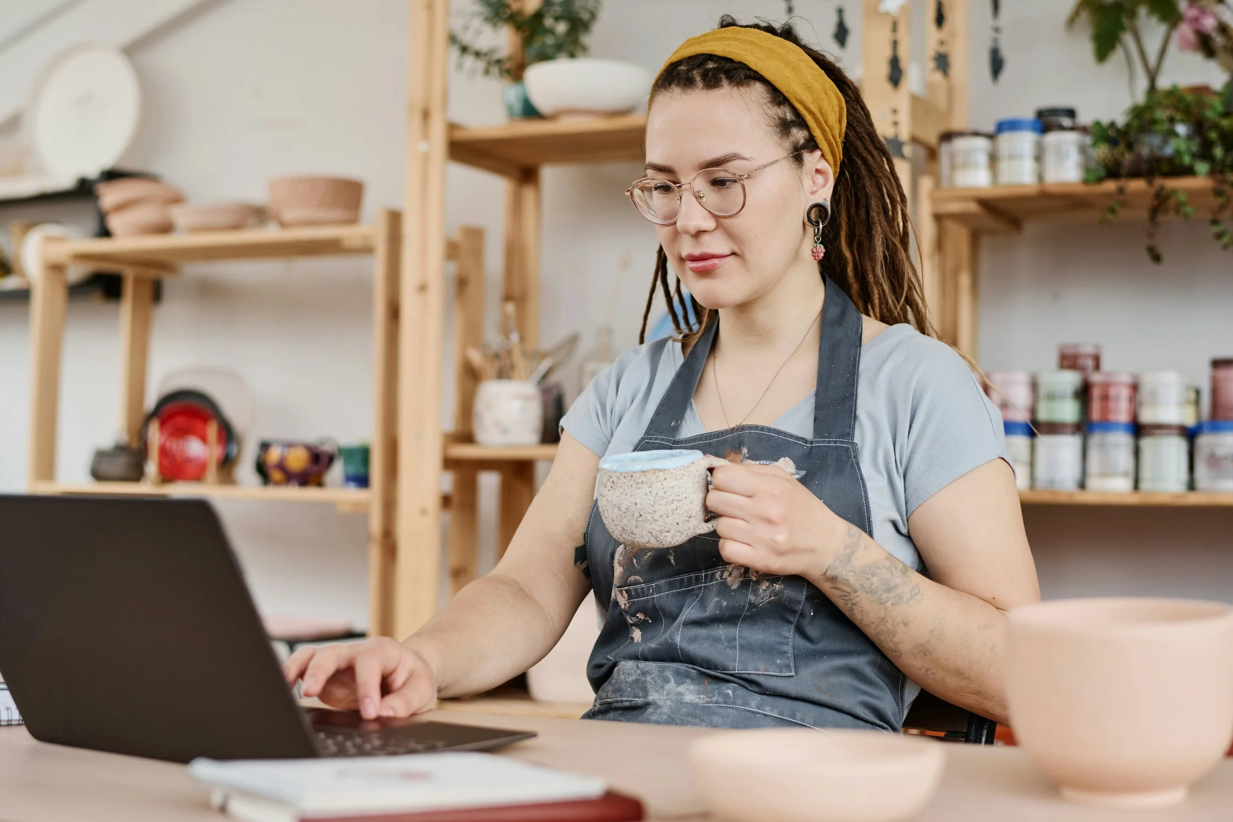 A woman with long dreadlocks, wearing glasses, a mustard headband, and a gray shirt, sitting at a table with a laptop, holding a mug in her right hand, in a pottery studio with shelves of pottery and supplies in the background.