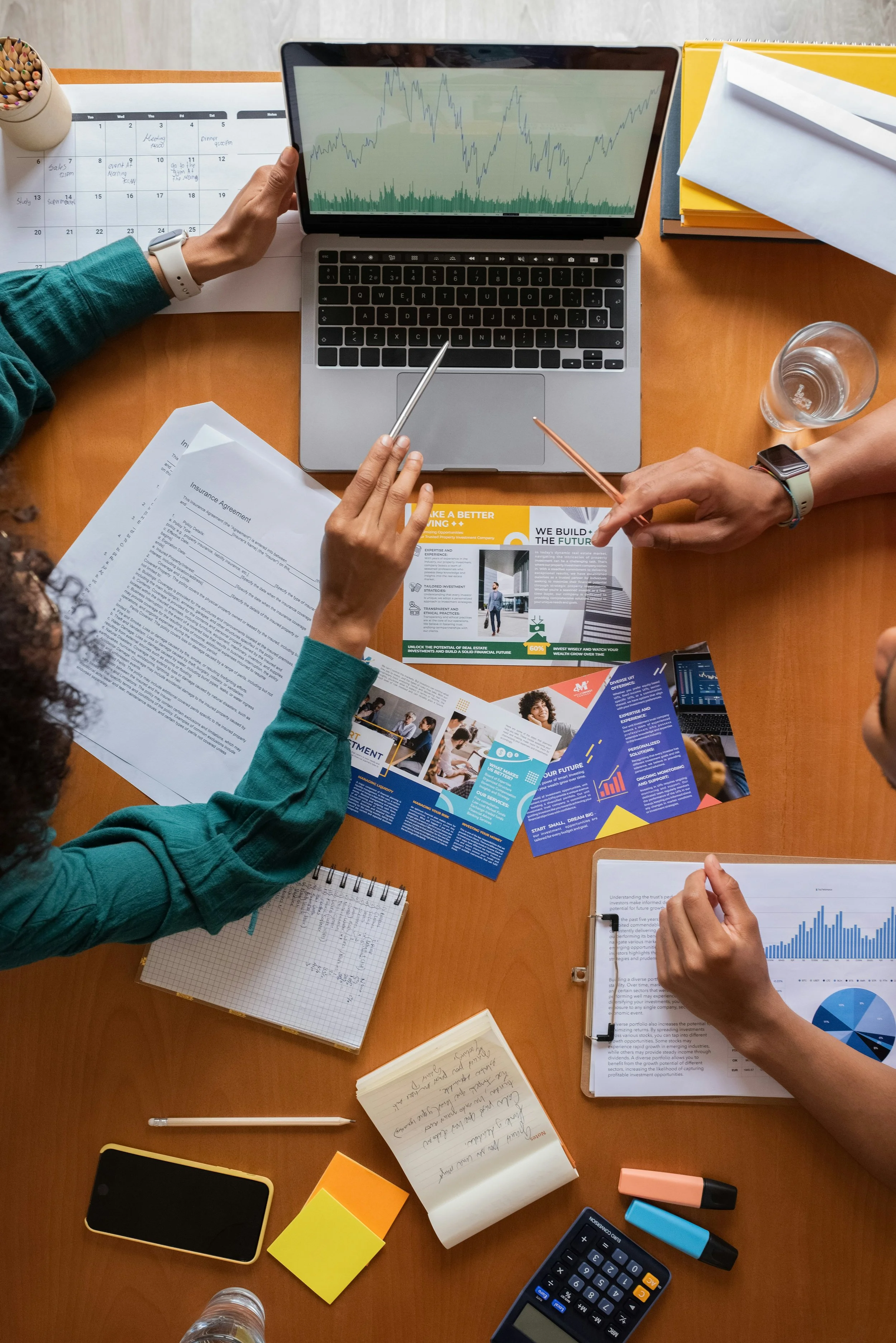 Overhead view of a group of people working on a business presentation at a wooden table, with documents, brochures, a laptop showing a graph, a tablet, a calculator, pens, sticky notes, and notebooks.