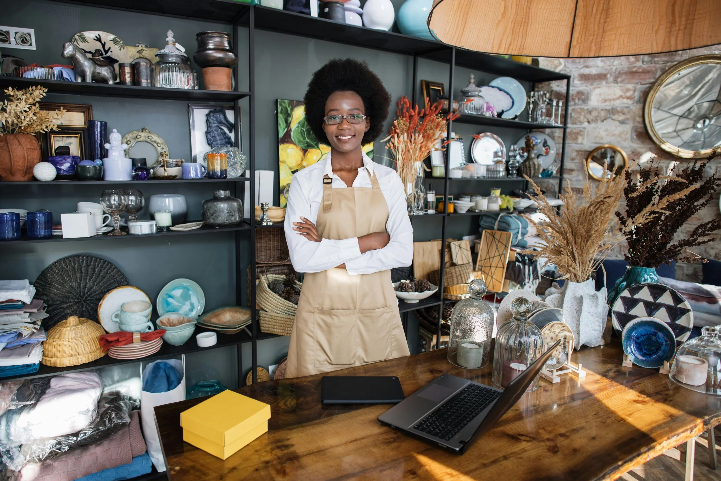 Woman with glasses and curly hair standing with arms crossed behind a wooden table in an antique shop or home decor store, surrounded by shelves of various decorative items, vases, and ceramics.