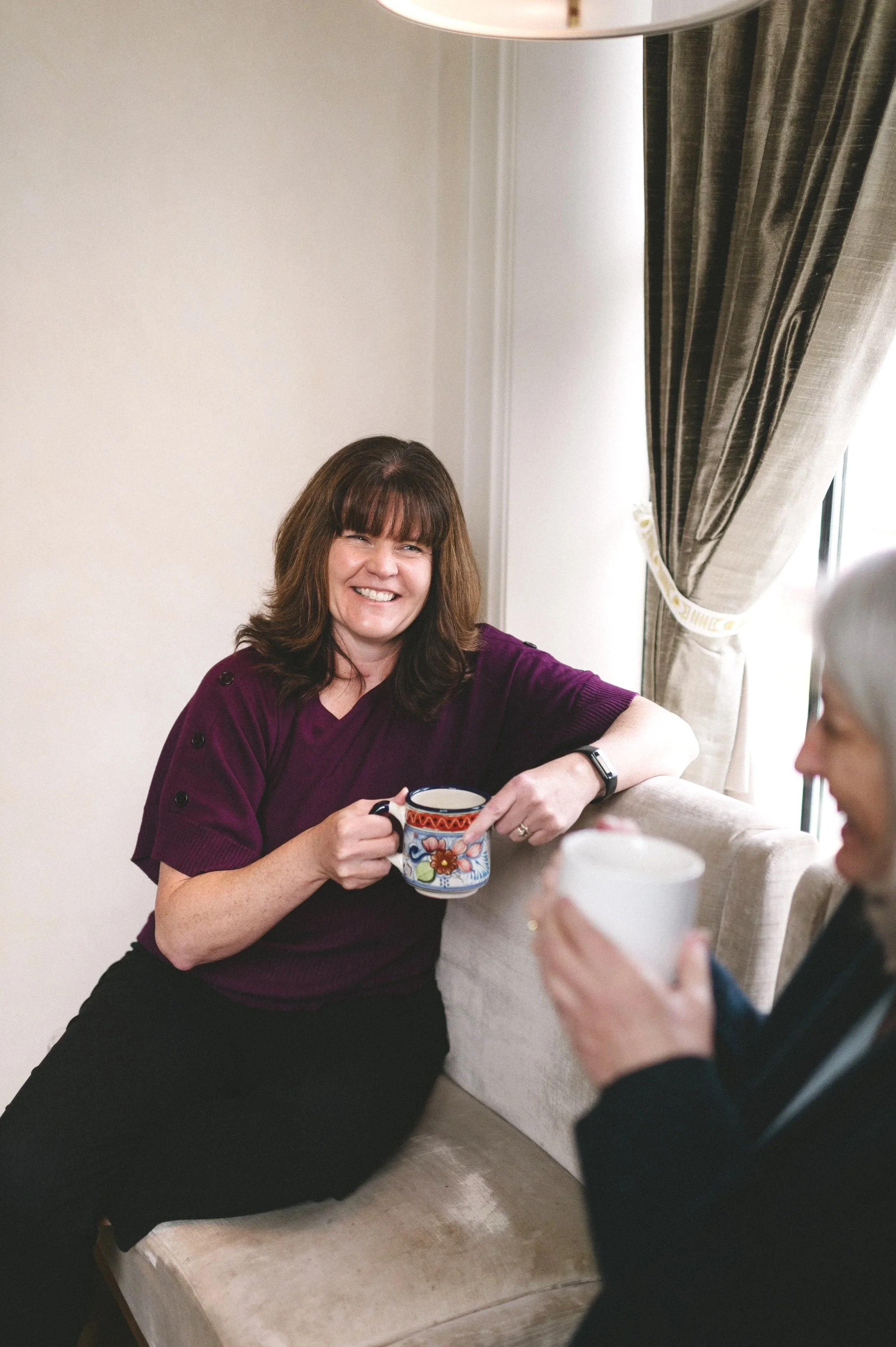 Crystal smiling and holding a colorful mug, sitting on a beige couch in a room with beige walls and a window with curtains. Another person with gray hair holding a mug is partially visible.