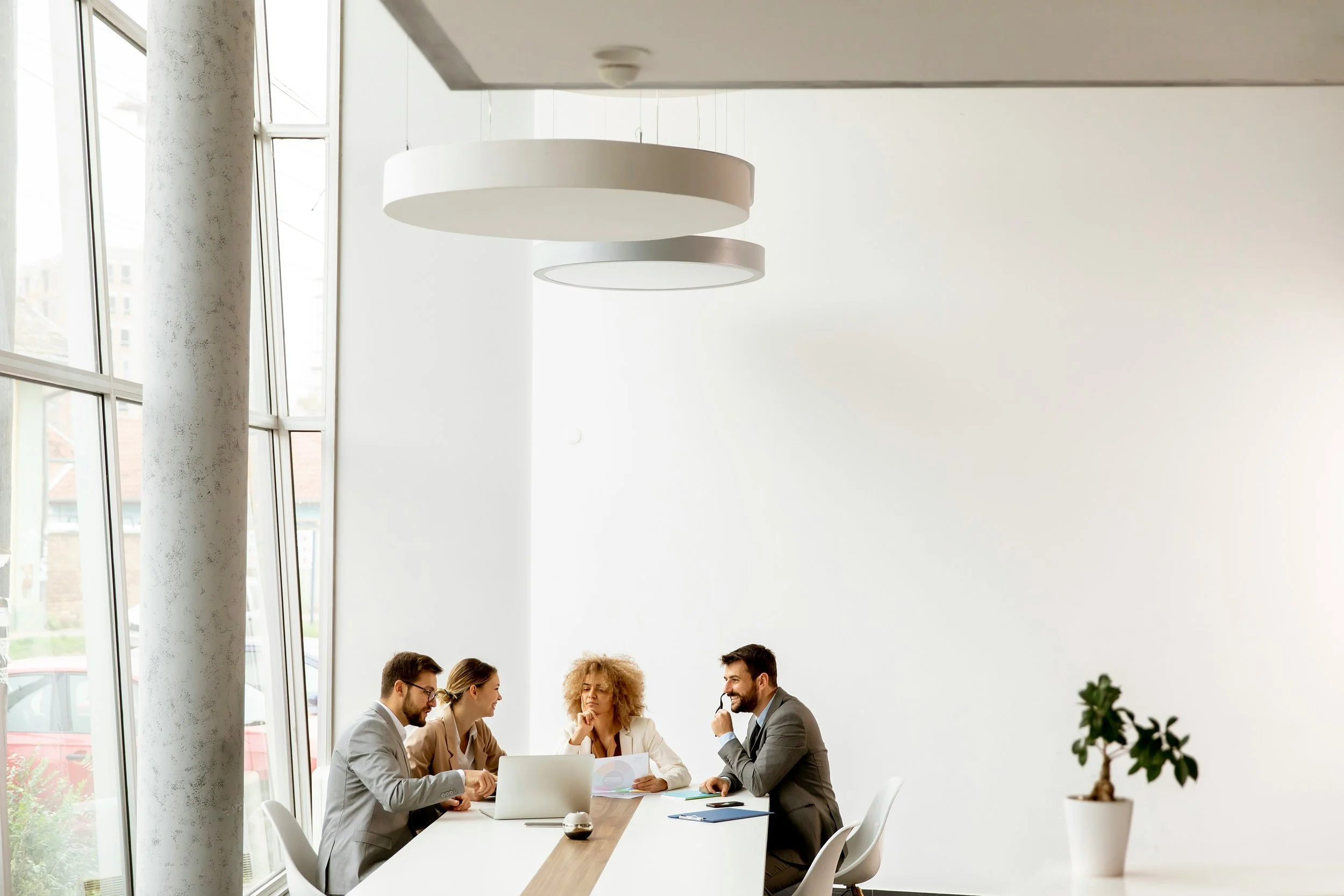 Four people in business attire having a meeting in a modern conference room with large windows, a white wall, and a potted plant.