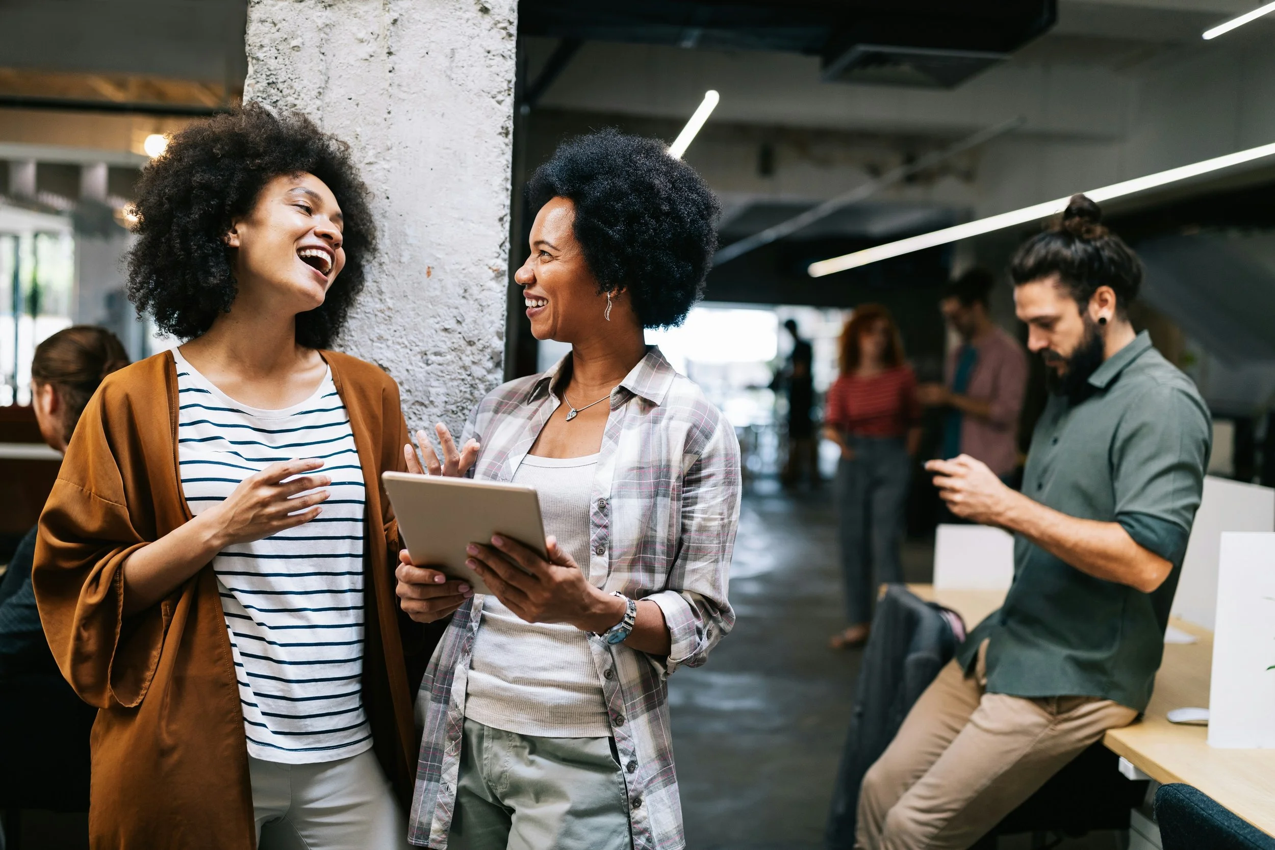 Two women engaging in conversation and smiling in a modern, open office space, with other people working in the background.