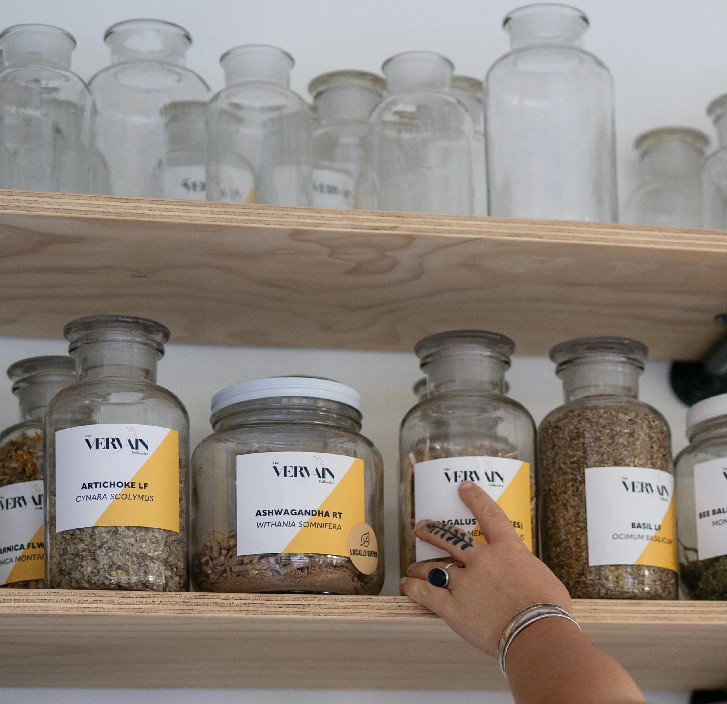 A hand reaching for a jar of herbs on a wooden shelf, with other glass jars of herbs and spices above and beside it.