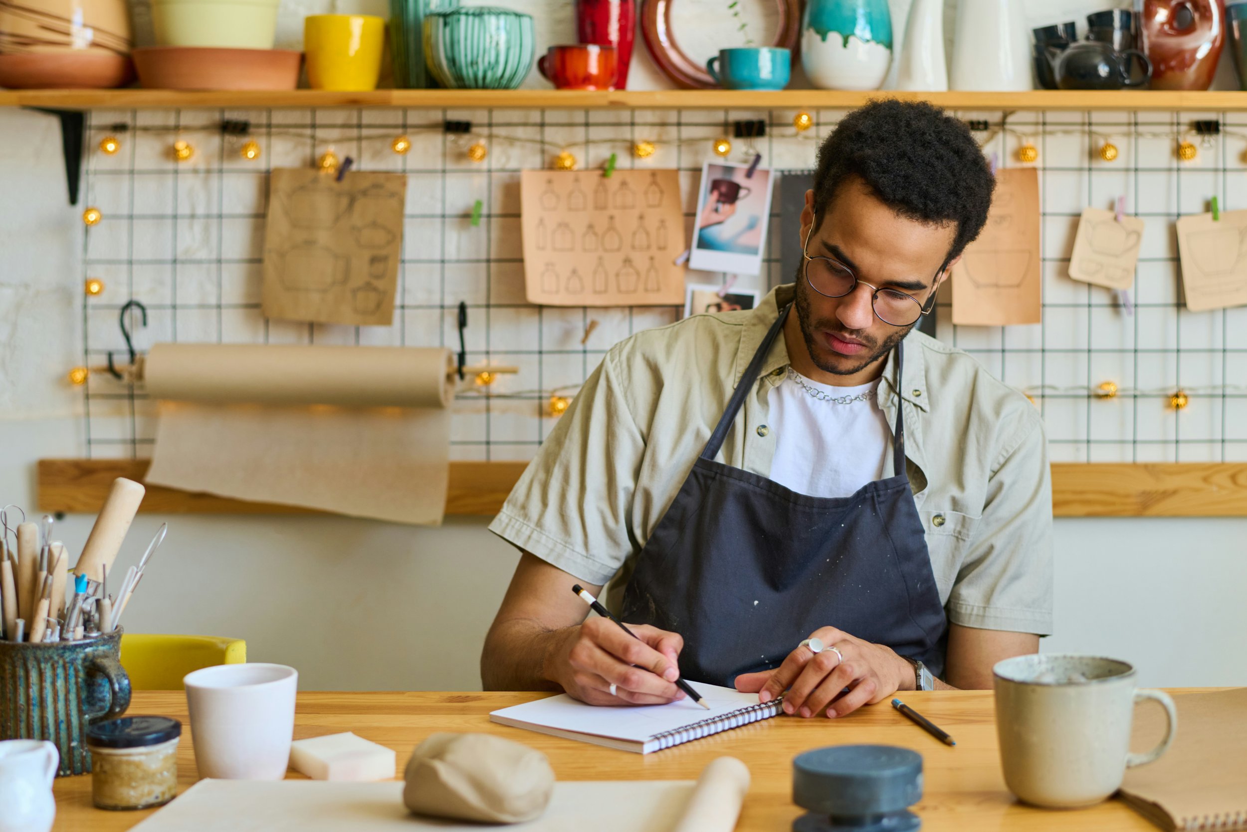A man wearing glasses and a black apron is sitting at a wooden table, drawing in a sketchbook, surrounded by art supplies. The background features a grid wall with sketches, photos, and hanging paper bags, and shelves with colorful pottery and cups.