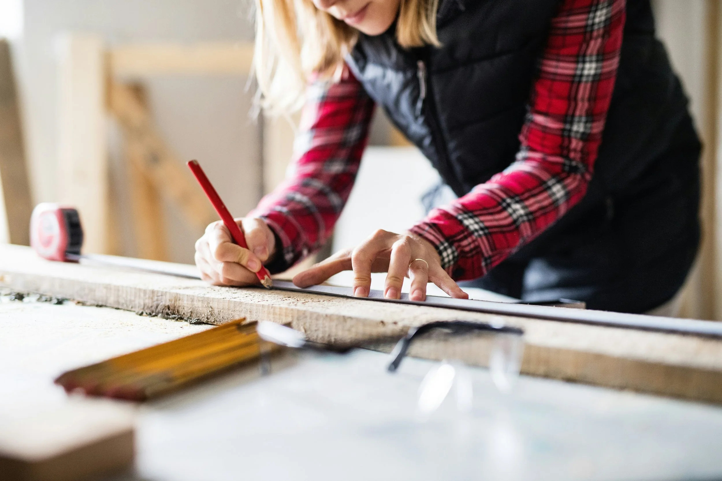 Person measuring and marking a piece of wood with a red pencil in a workshop.