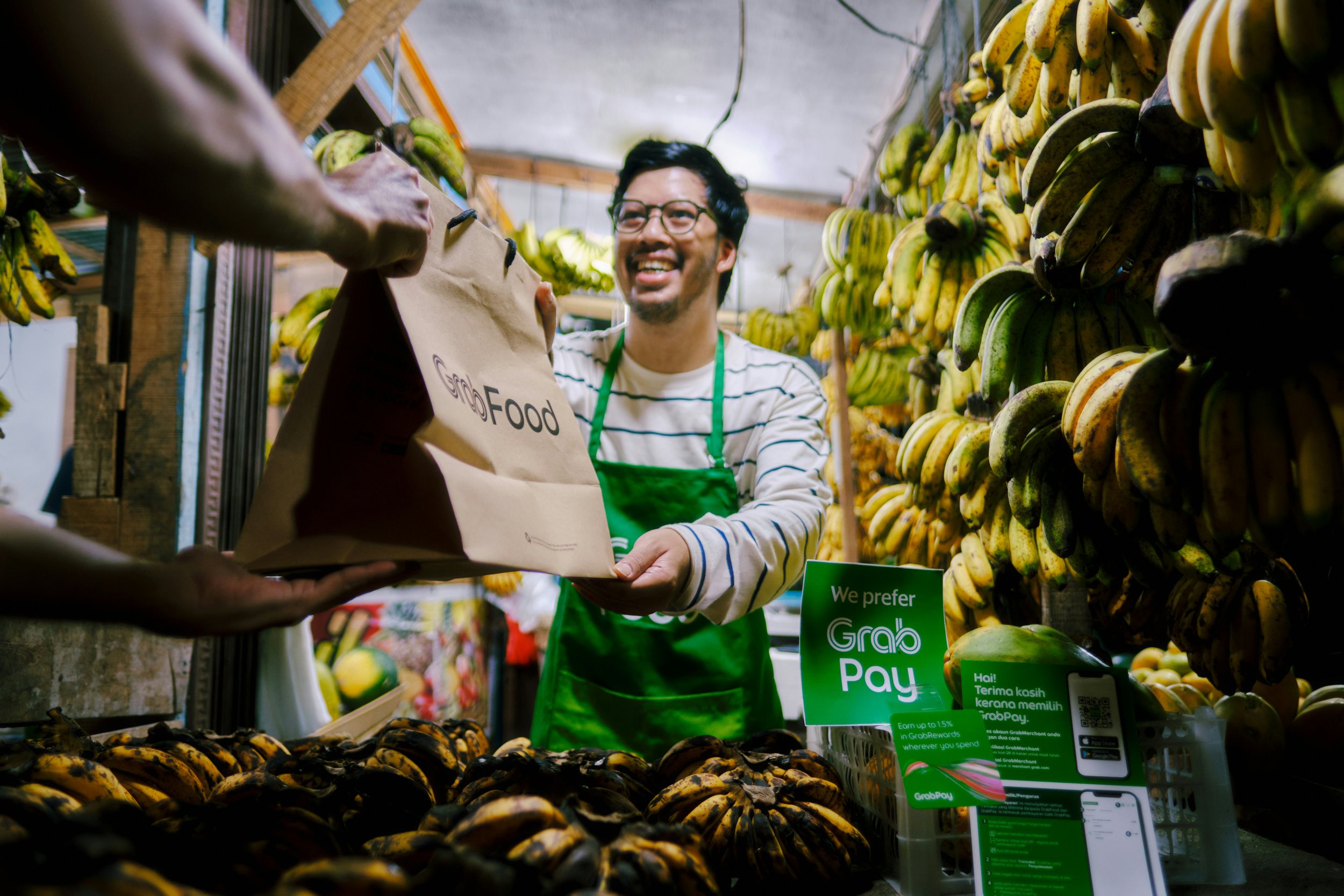 A man in a striped shirt and green apron smiling as he hands a paper bag to a customer at a banana stand, surrounded by bunches of ripe bananas, with a sign indicating preference for GrabPay.