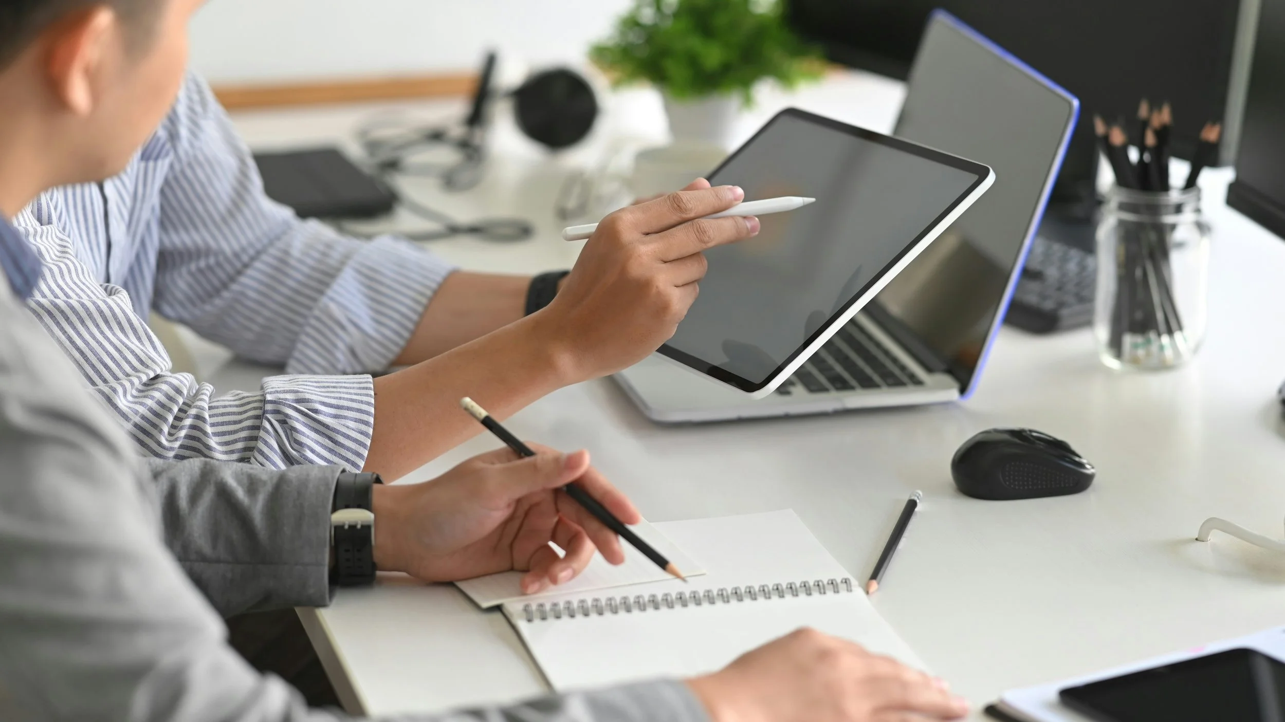 Two people working at a white desk with a tablet, laptop, notebook, and office supplies, in an office environment.