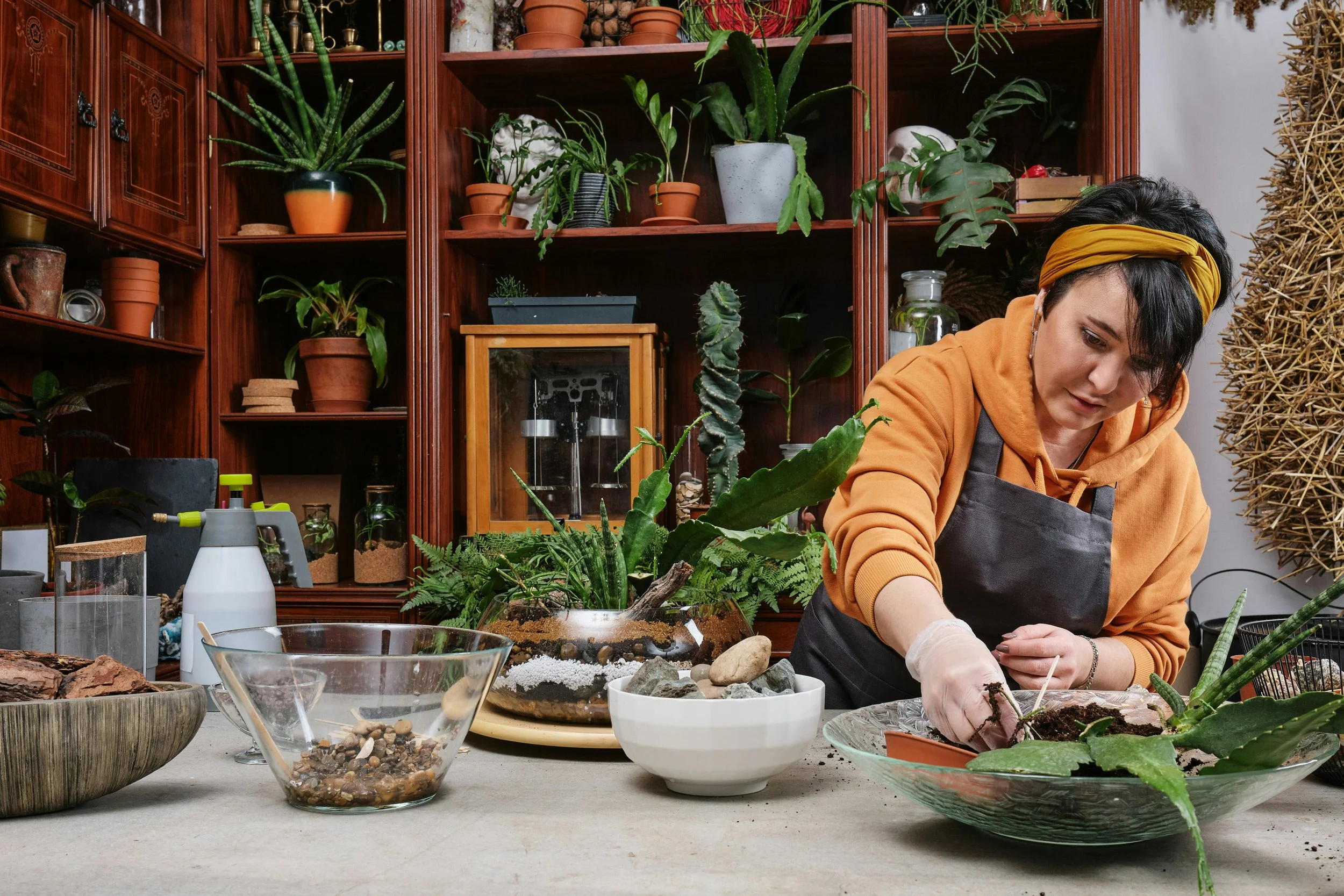 A woman wearing an orange hoodie, black apron, and yellow headband is potting a plant in a glass bowl. The workspace is filled with various plants, gardening tools, rocks, and soil, with a wooden shelf filled with potted plants in the background.