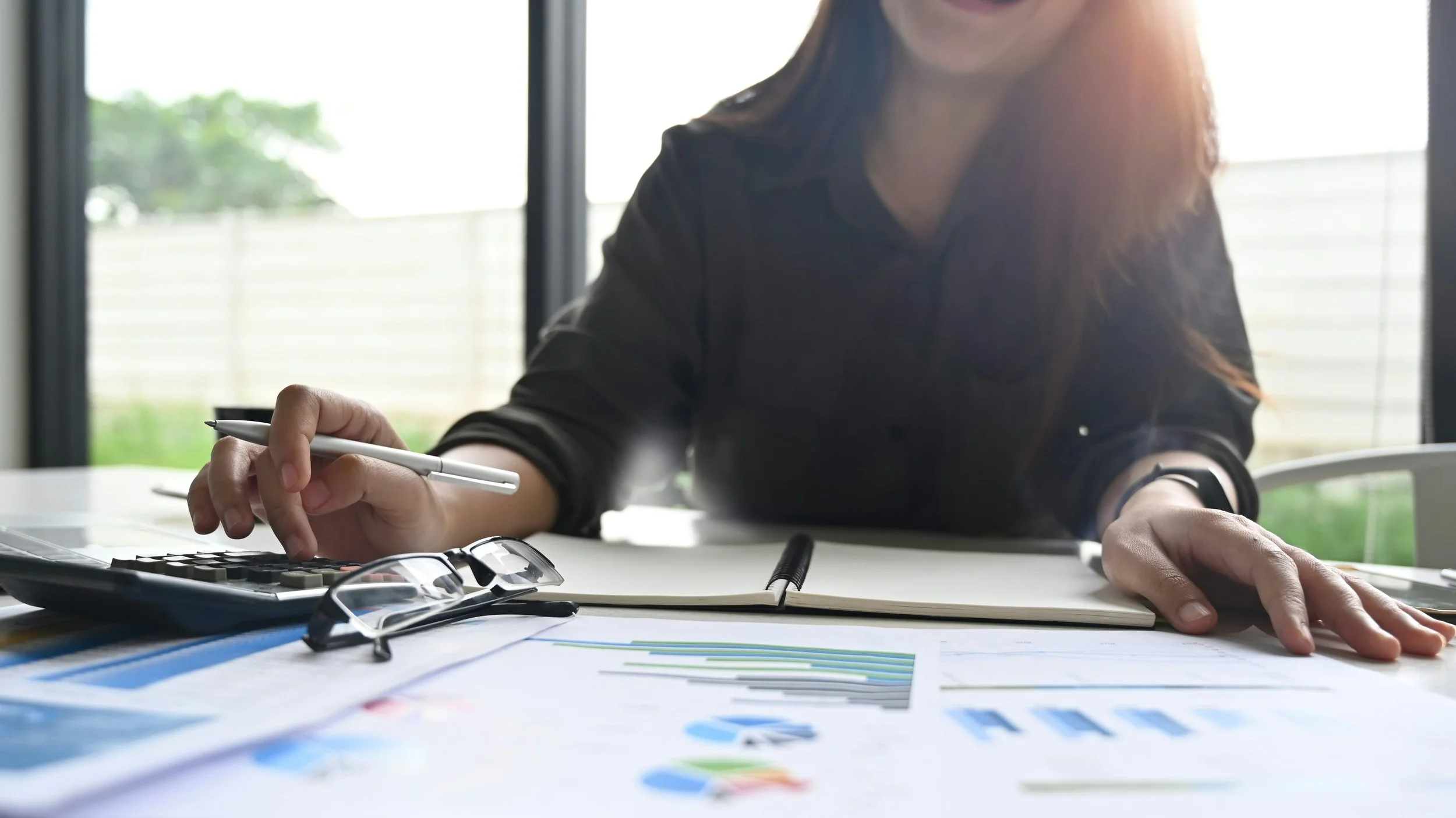 A woman sitting at a desk with documents, a calculator, and glasses, working with a pen, during daytime.