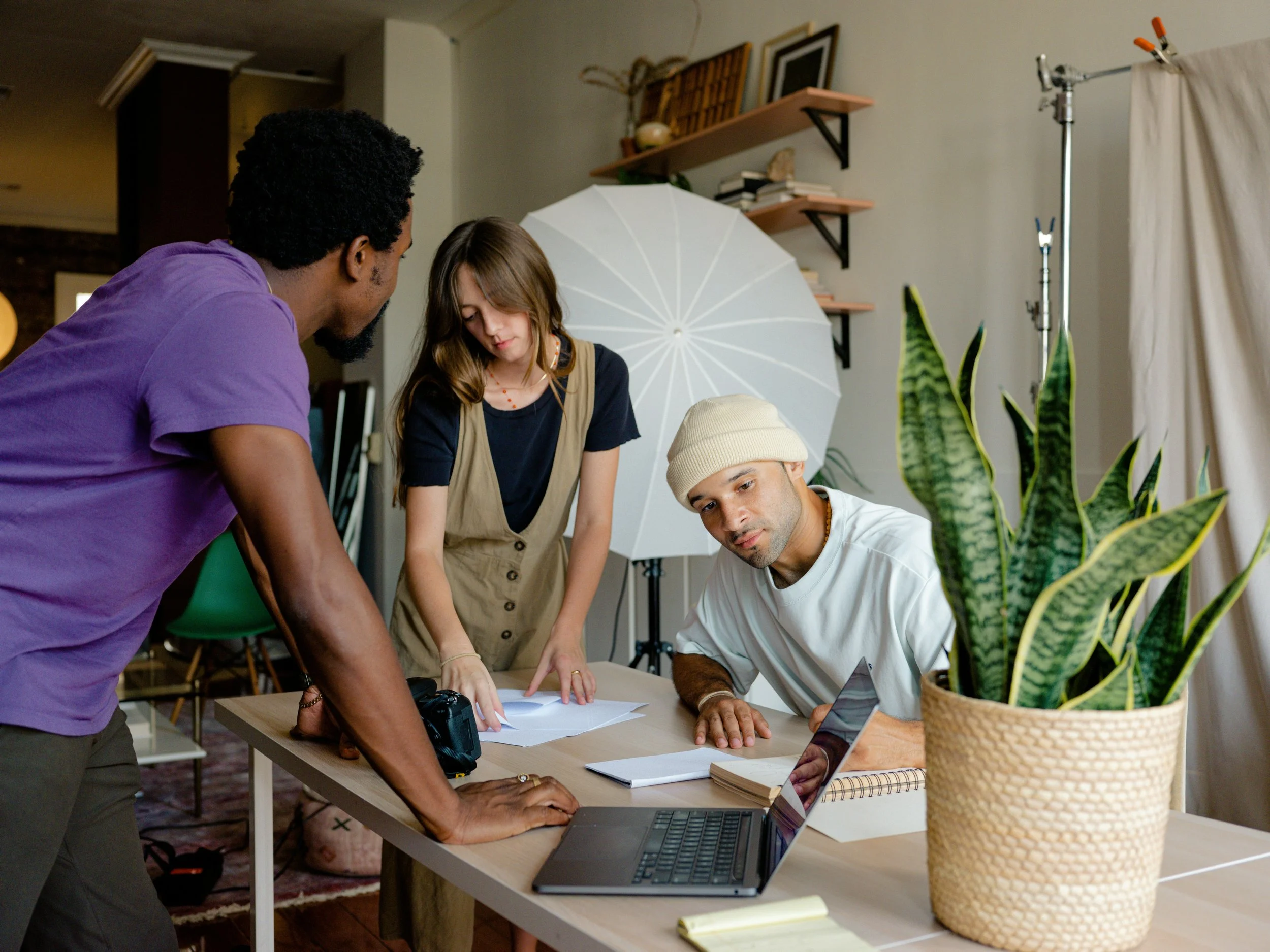 Three people collaborating around a table in a photography studio with a large white umbrella, books, a laptop, and a potted plant.