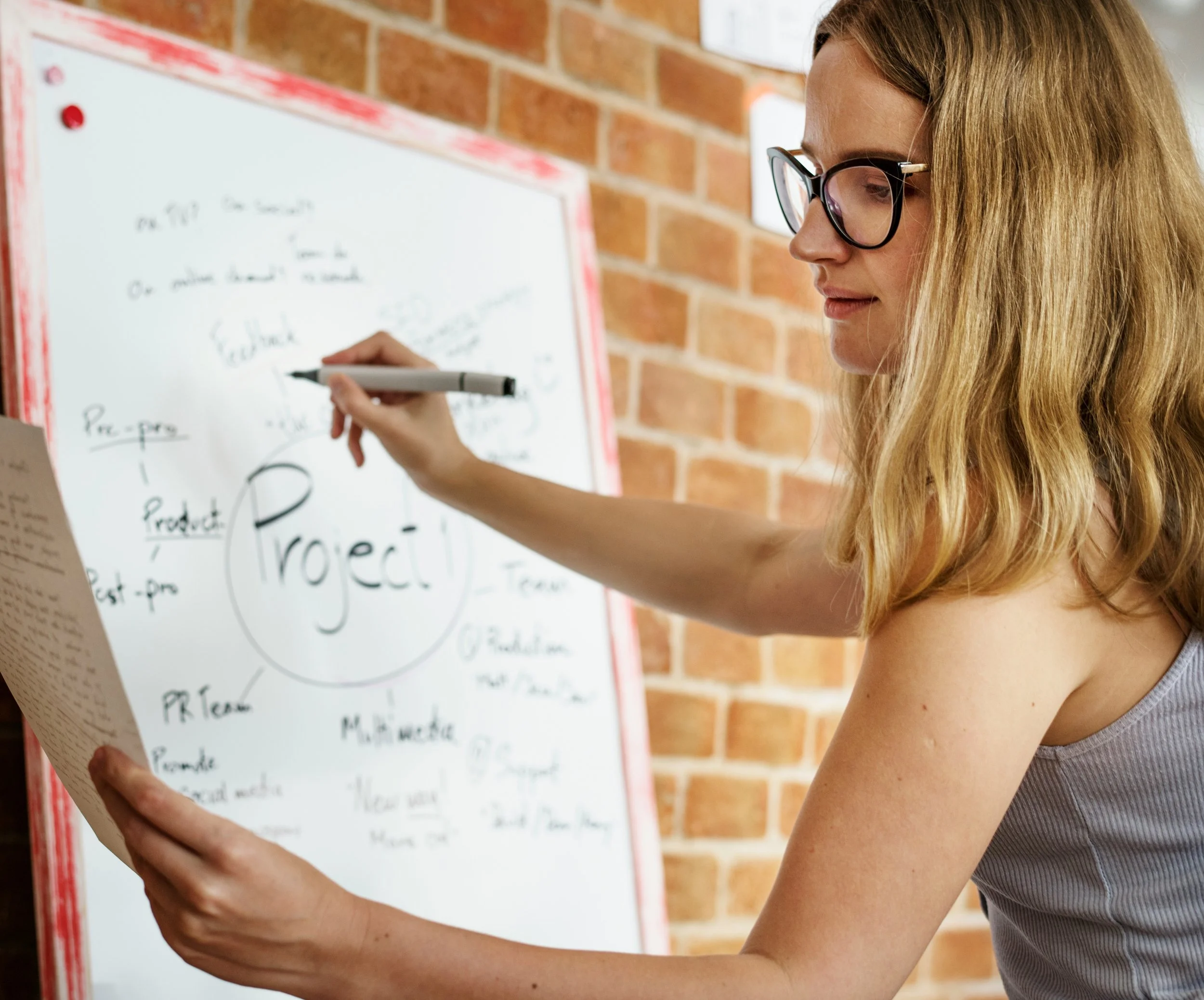 A woman wearing glasses and a sleeveless top writes on a whiteboard with a black marker, with a brick wall in the background.