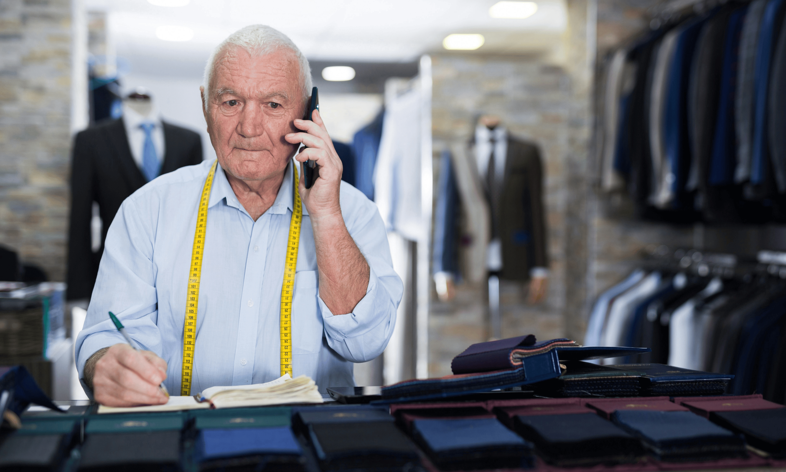An elderly man with white hair wearing a light blue shirt and a yellow measuring tape around his neck, talking on a cellphone inside a men's clothing store, with fabric swatches and notebooks on the counter in front of him.