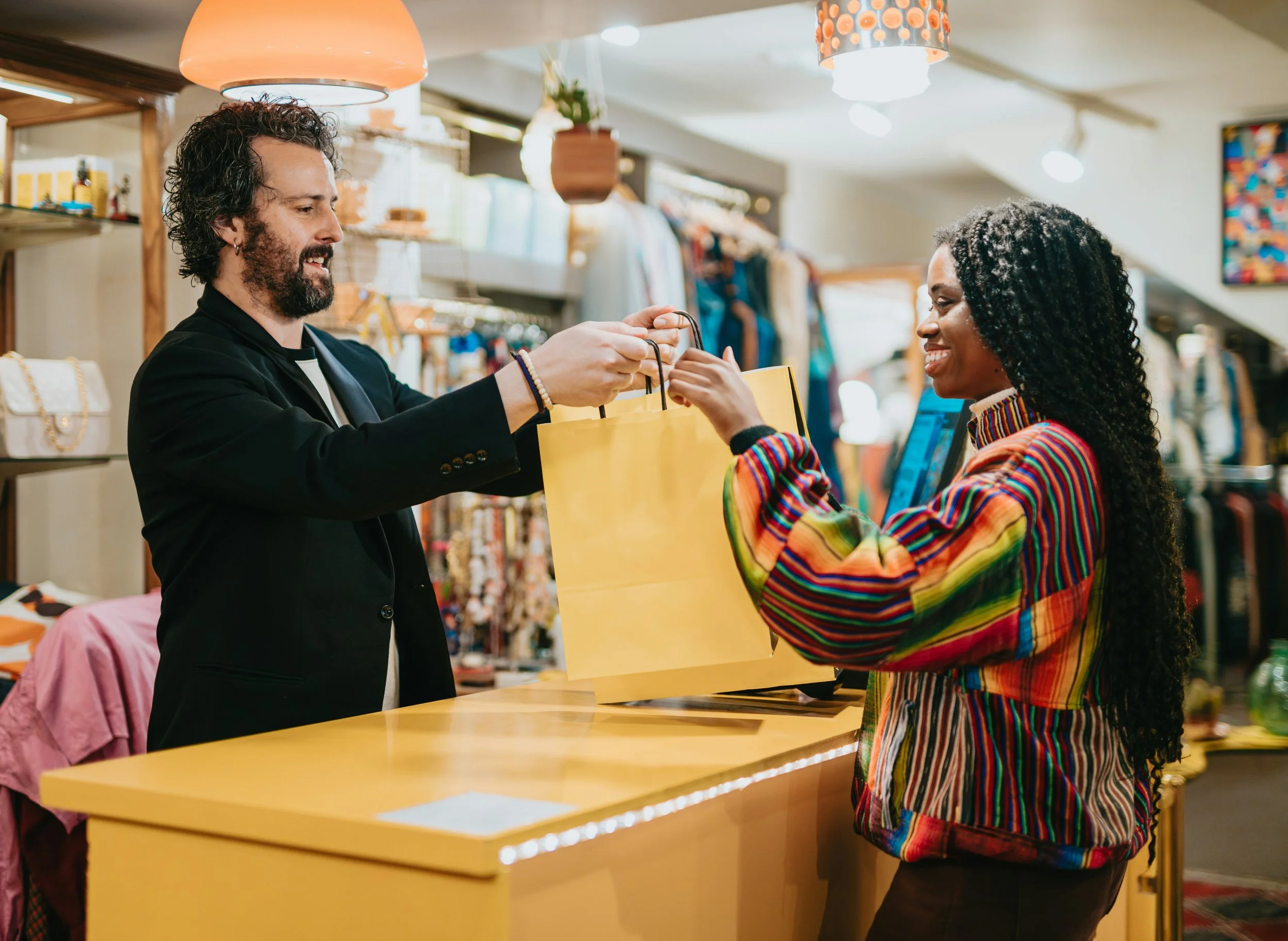 A man with curly hair and a beard hands a yellow shopping bag to a woman with long curly hair wearing a multicolored striped jacket at a retail store counter. The woman is smiling as she receives the bag.