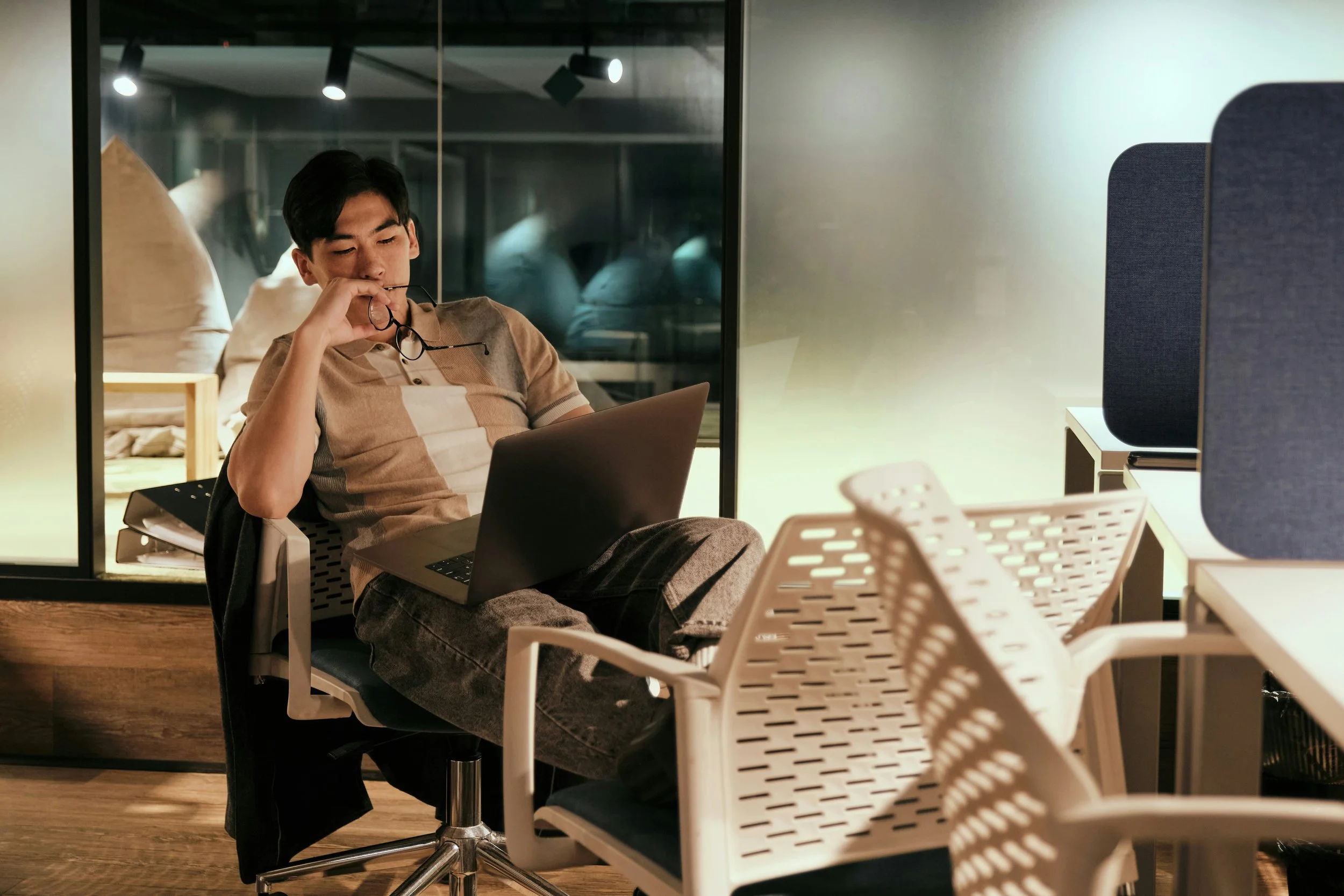 A young man sitting at a desk in an office, looking at a laptop and holding his glasses.