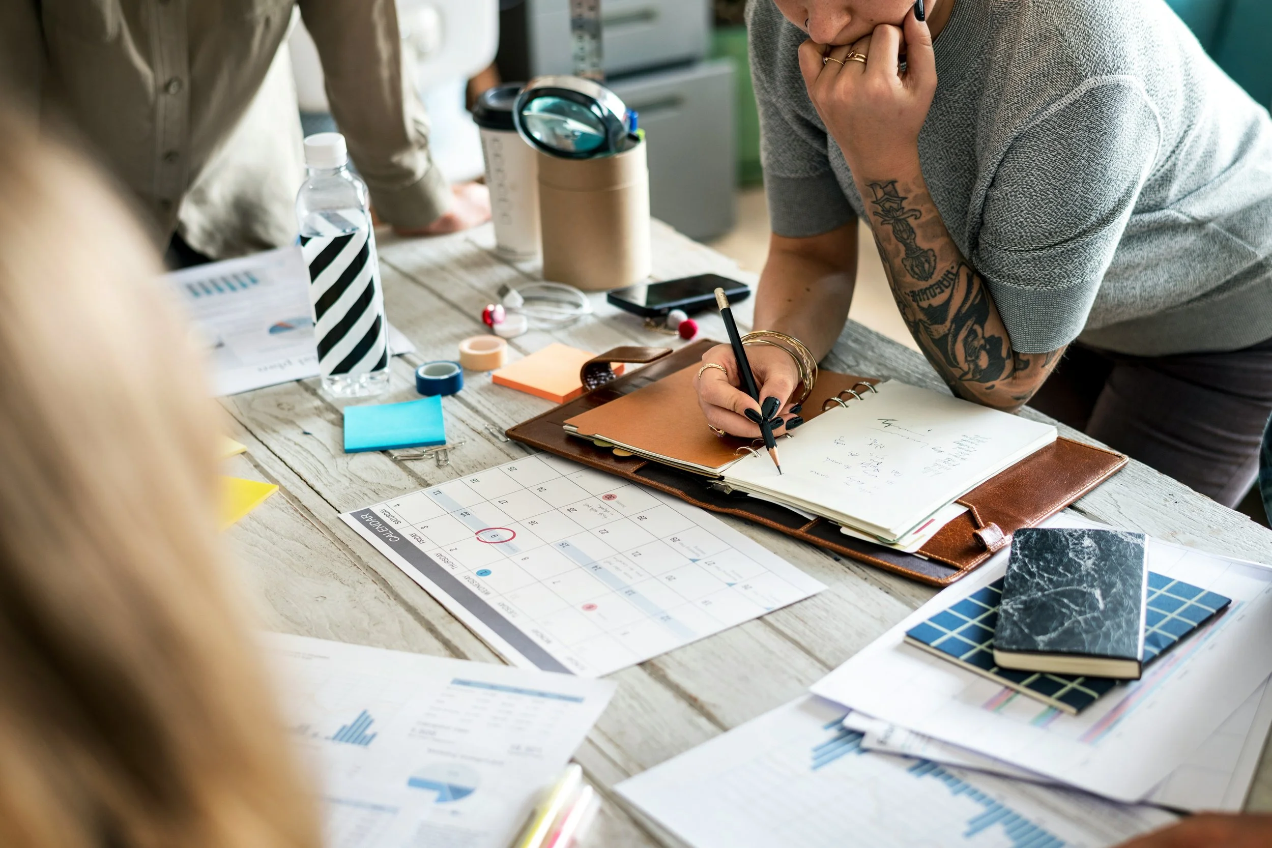 A woman with tattoos is writing in a notebook on a cluttered desk during a meeting.