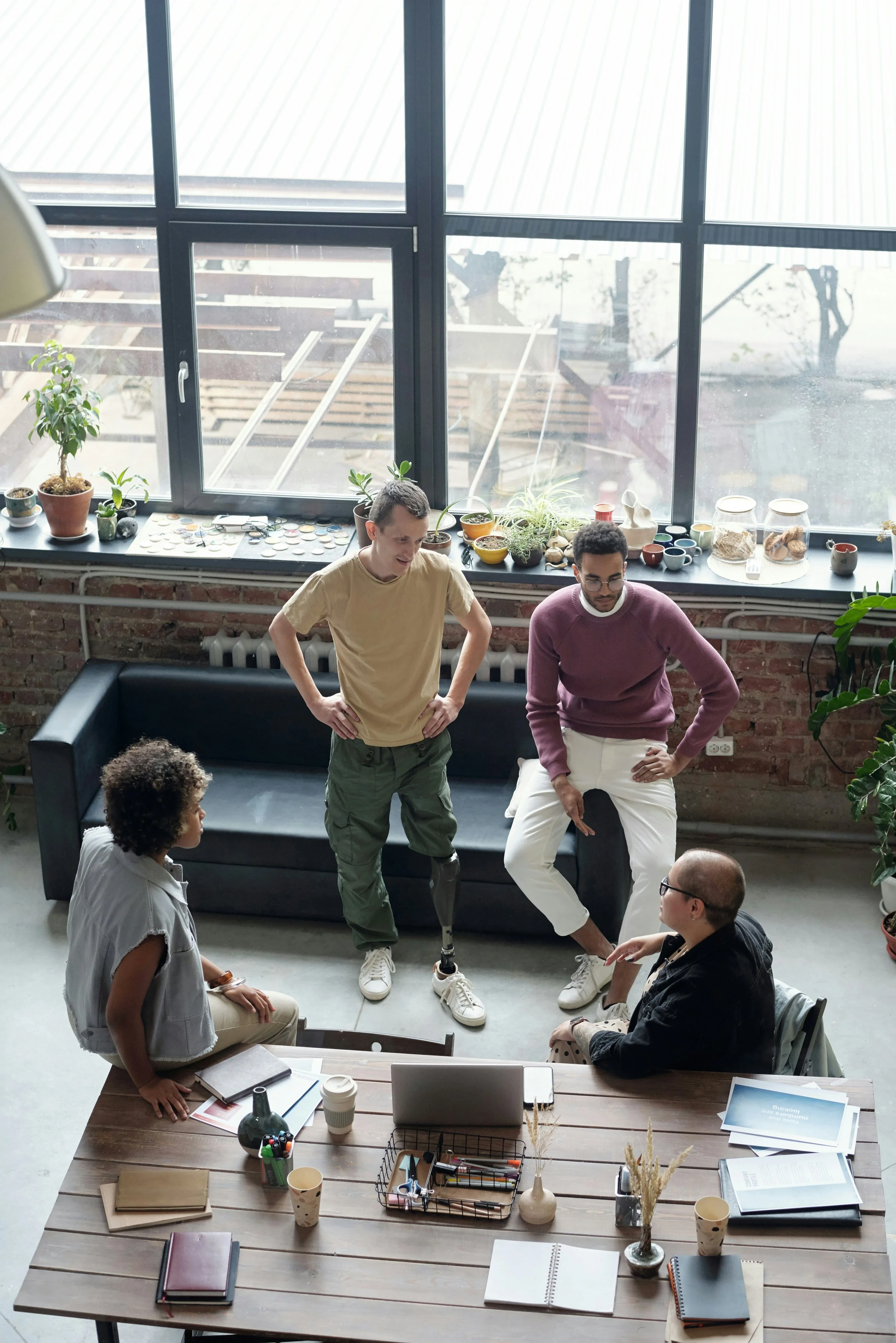 Group of five people having a discussion in a modern co-working space with large windows, potted plants, and a wooden table with notebooks, pens, and a laptop.