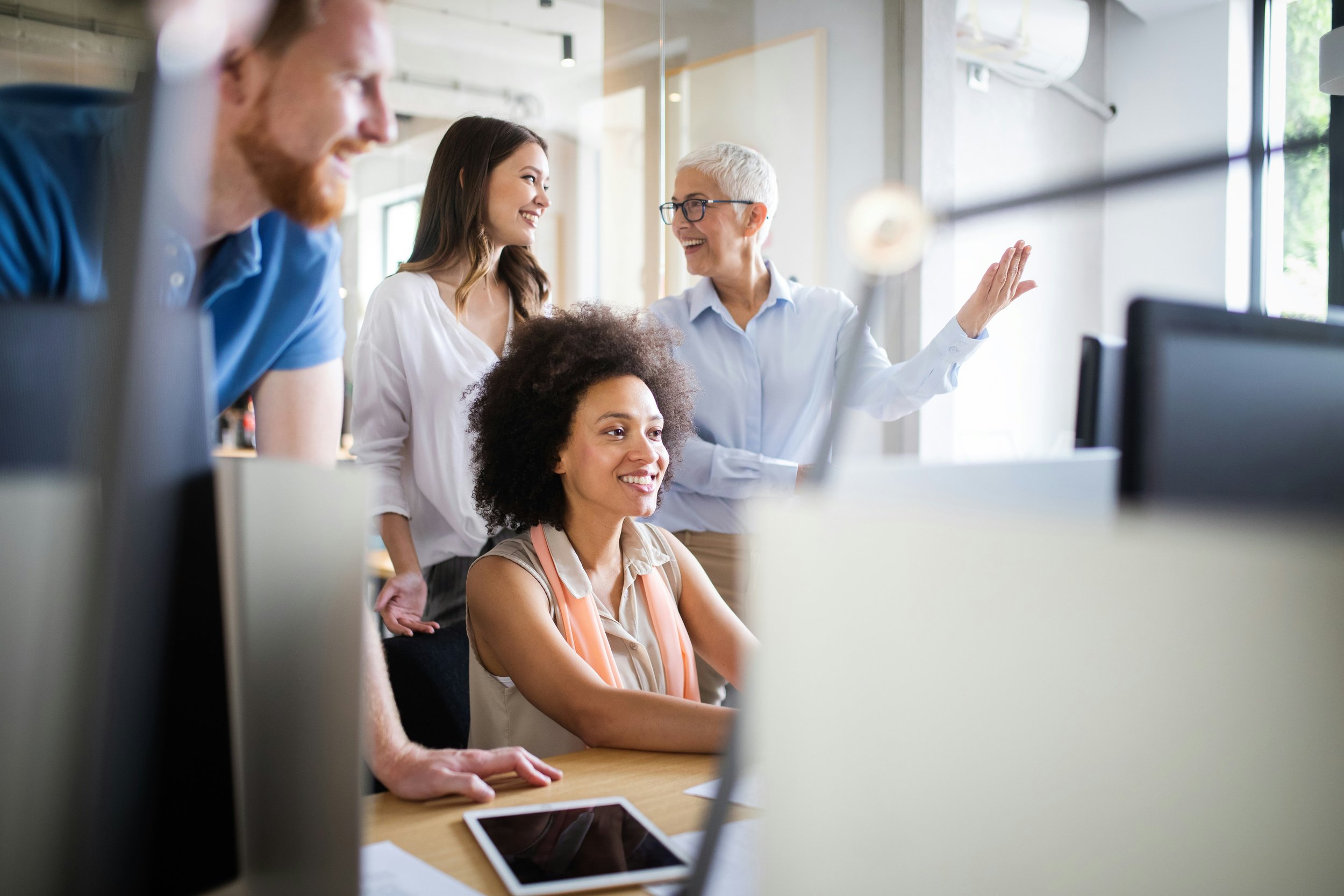 A group of five women in a bright office space, engaged in a discussion or presentation, with one woman standing and gesturing while the others are smiling and listening.