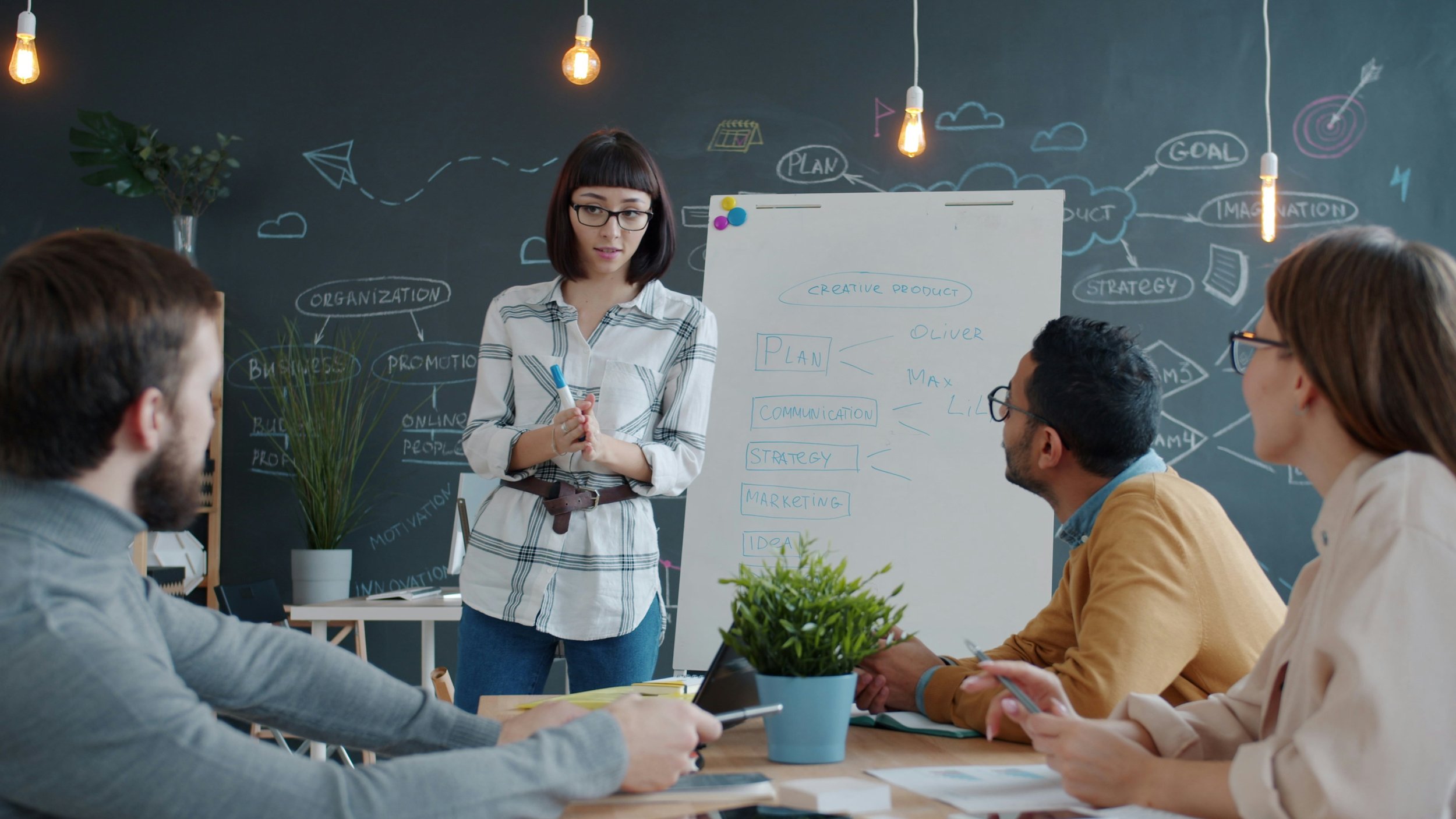 A diverse group of four people in a meeting room with a blackboard wall covered in doodles and notes. A woman in glasses and a white plaid shirt stands near a whiteboard, presenting to three seated colleagues, with plants and hanging lights in the room.