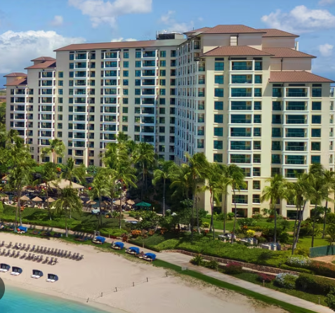 A large beachfront hotel with white exterior and multiple floors, surrounded by palm trees and a sandy beach.