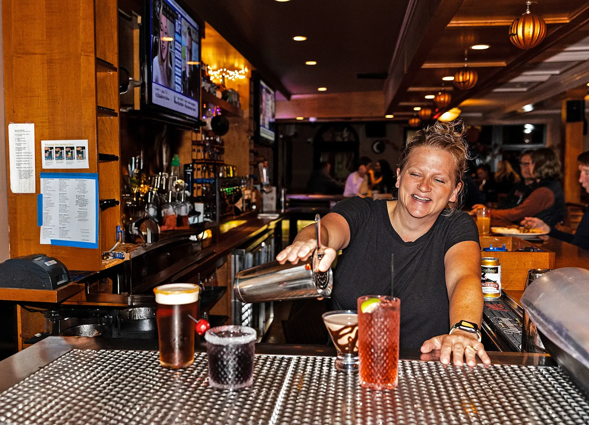 A woman smiling and pouring a drink at a bar. The bar has various bottles and glasses, and multiple drinks on the counter. There are several TVs and people in the background.