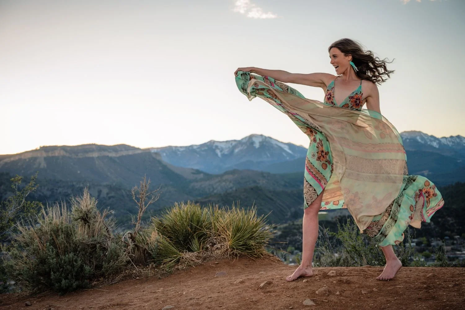 A woman in a colorful, flowing dress is standing barefoot on a dirt hilltop, holding her dress out as it flows in the wind. She is smiling and looking to her left with mountains in the background during sunset or sunrise.