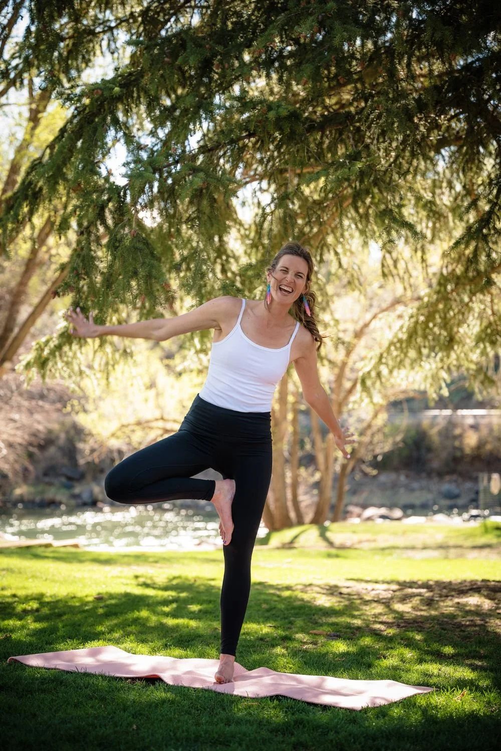 A woman practicing yoga outdoors on a pink mat under a tree, smiling and balancing in the tree pose.