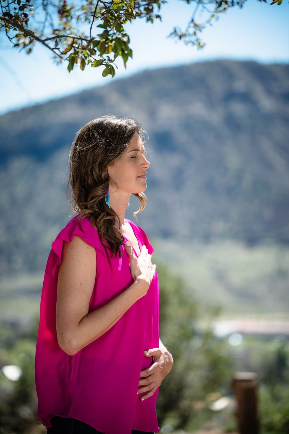 A pregnant woman in a bright pink top with ruffled sleeves, standing outdoors with her eyes closed and her right hand over her heart, left hand on her belly, near mountains and trees under a clear blue sky.