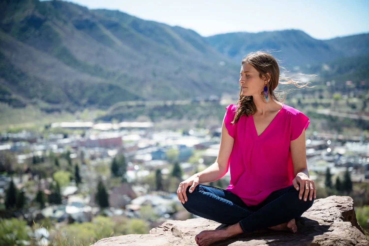 A woman sitting cross-legged on a rock in a meditative pose outdoors with mountains and a town in the background, wearing a bright pink top and dark jeans.